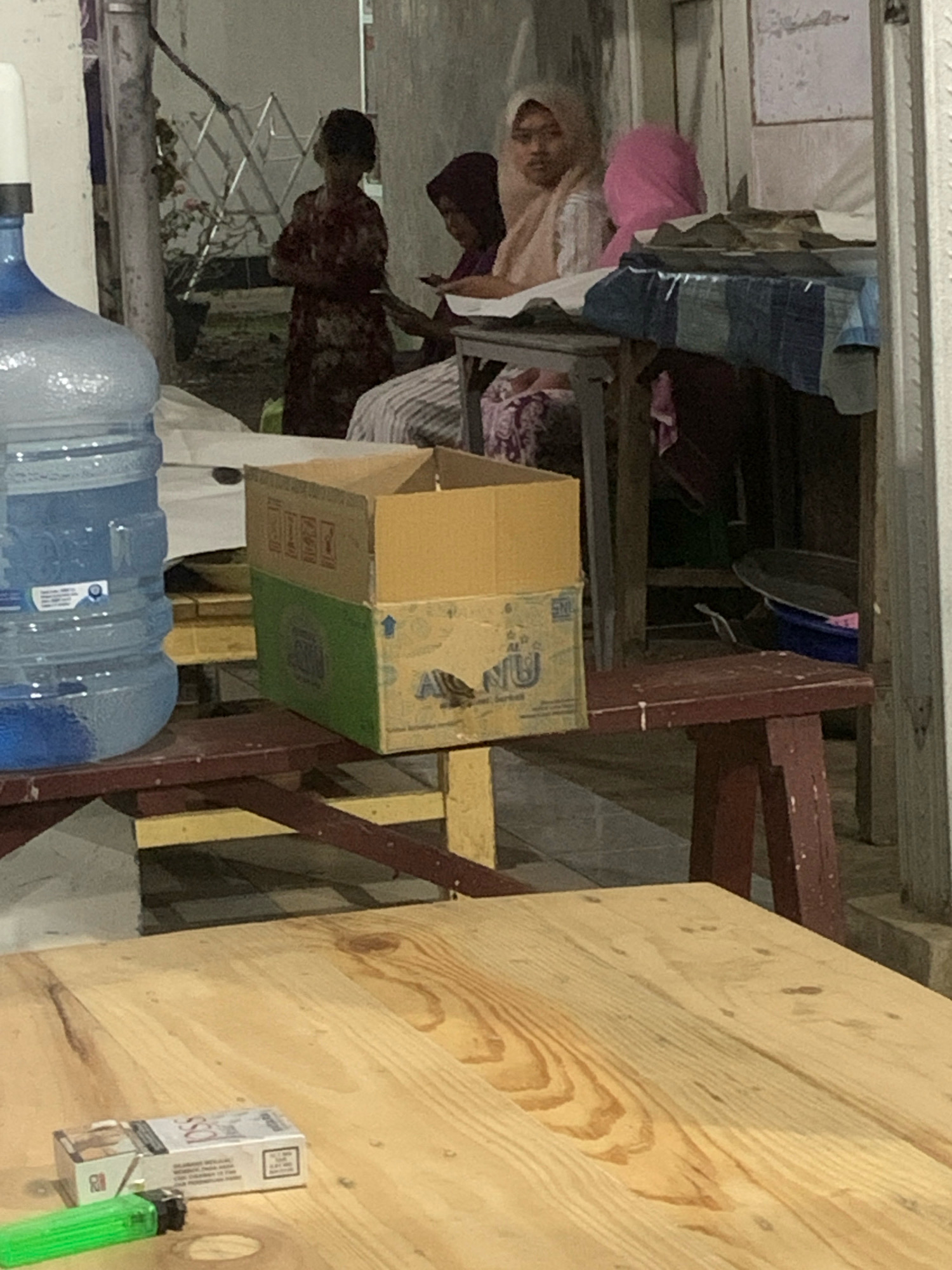 A group of individuals, including at least two women wearing hijabs, are gathered around a table outside a building. One person is standing, wearing a patterned dress. On a table in the foreground, a pack of cigarettes and a green lighter are visible. A large blue water jug is positioned to the left. A cardboard box is stacked on top of another box. The environment appears to be a street scene in Bululawang, Indonesia, possibly an informal eatery or gathering space, with elements of both indoor and outdoor settings.