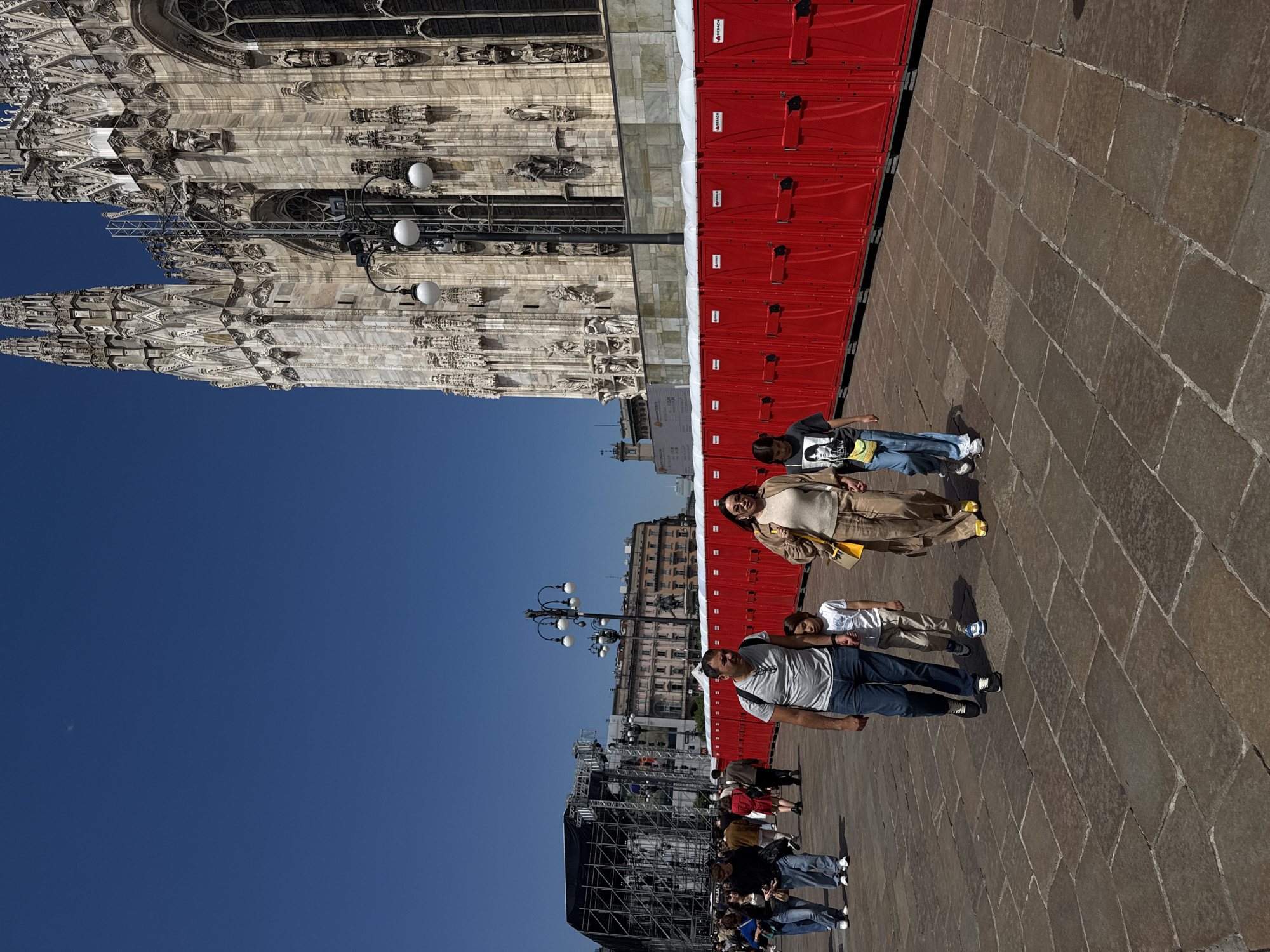 The image depicts a partially obscured facade of the Milan Cathedral (Duomo di Milano) and a plaza area. A long row of red portable toilets stands adjacent to the Cathedral. A group of people is walking along the paved plaza, including a man, a child, and a woman. The sky is clear and blue. In the distance, buildings and scaffolding can be seen. The image is rotated, positioning the cathedral on its side.