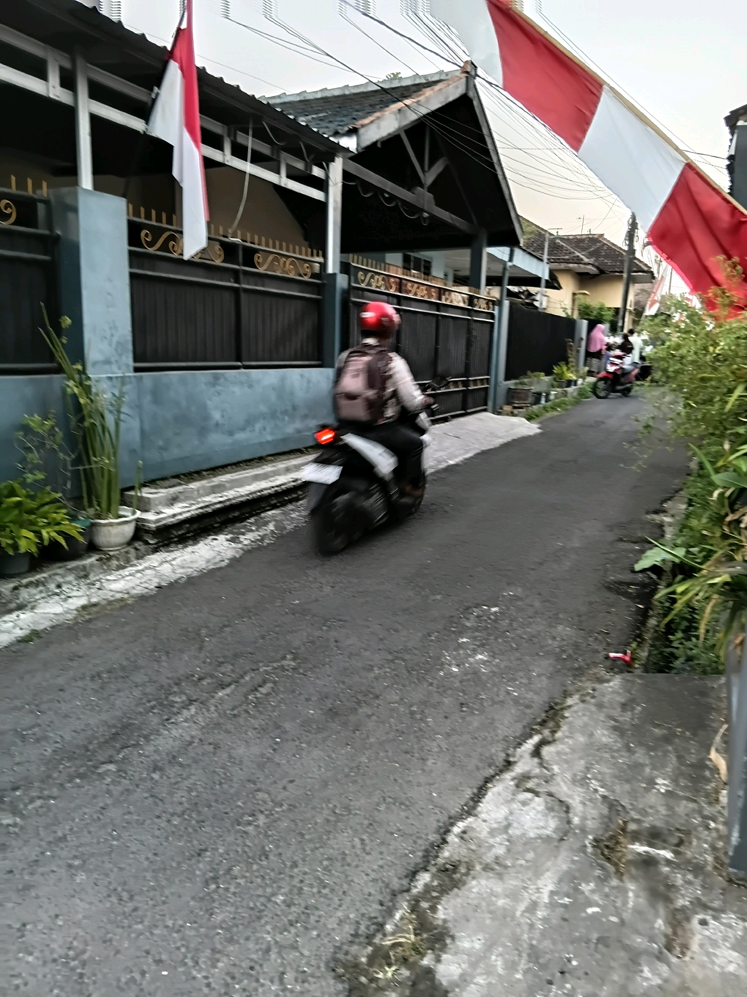 In Kebonsari, Indonesia, a residential street is depicted under an overcast sky. A motorcycle rider wearing a red helmet and a backpack is moving away from the camera down the asphalt road. The rider is positioned in the left half of the frame, with the motorcycle's rear visible. To the left of the road, a dark gray fence runs along a building with a covered porch. Potted plants are situated in front of the fence. Large red and white Indonesian flags are prominently displayed, stretching across the upper portion of the image from poles attached to the building. In the distance, on the right side of the road, another motorcycle is visible with two people on it, also moving away from the camera. Lush green foliage lines the right side of the street. The ground surface of the road shows some wear and tear, with cracks and a white painted line marking the edge of the road on the left.
