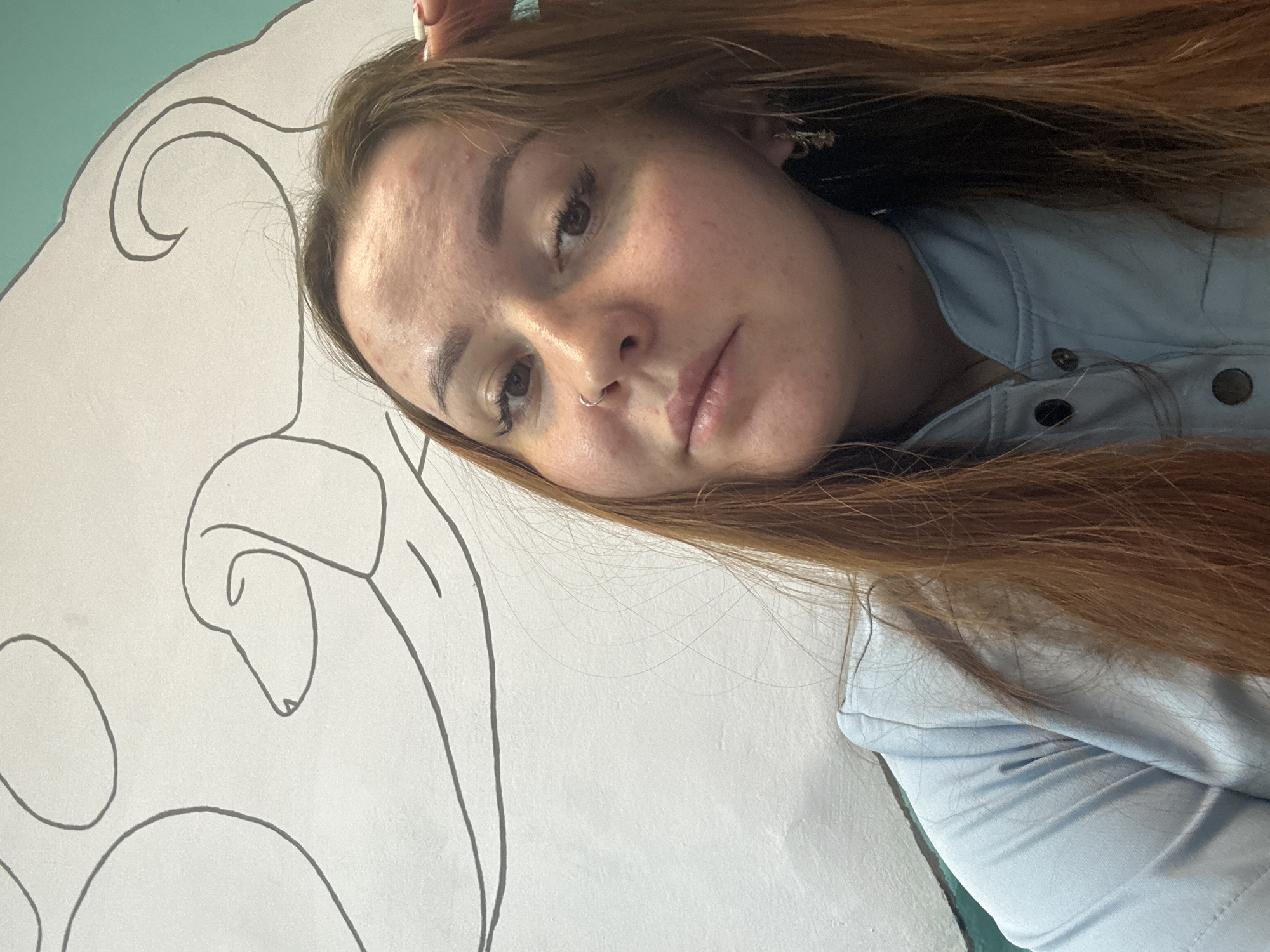 A young woman with long, reddish-brown hair lies down, looking directly at the camera with a neutral expression. Her head is tilted to the right, and her hair is spread around her. She wears a light blue top or jacket with dark buttons. Her face shows some minor skin imperfections, and she has a small silver hoop piercing in her left nostril. Her eyes are open, and her eyelashes appear prominent.
The background consists of a light-colored wall with stylized black outline drawings, possibly depicting clouds or abstract shapes, one of which has a flowing, curled top portion and another resembling a dog or mythical creature's head. Part of the wall, visible in the upper left, is painted a light teal or turquoise color. The image is framed as a close-up, focusing primarily on the woman and a significant portion of the illustrated wall. There is no visible text. The activity appears to be posing for a photograph indoors in Izhevsk, Russia.