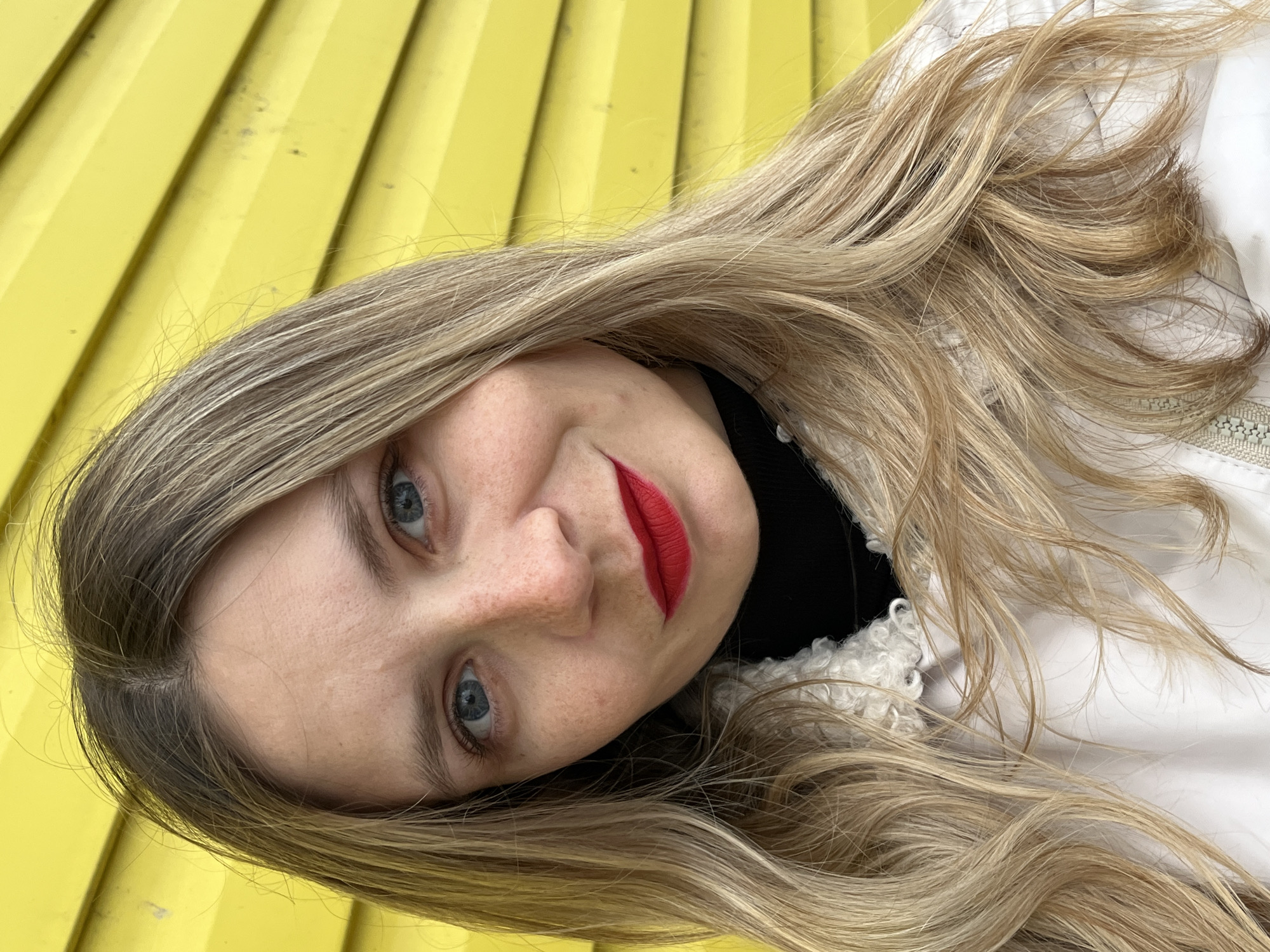 A close-up, slightly angled shot of a light-skinned woman with blonde hair, blue eyes, and red lipstick. She is wearing a black turtleneck sweater and a white jacket. The background is a corrugated yellow wall. The location is likely Chaikovsky, Russia, based on available data.