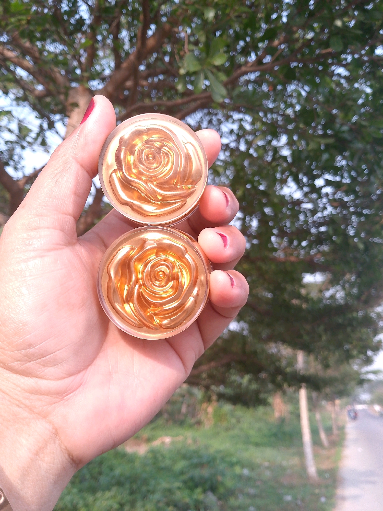 In a medium shot, a hand with manicured fingernails holds two small, circular, rose-shaped objects. The objects are gold in color and appear to be made of a shiny material. The background is blurred, showing green foliage and a hint of a road with a vehicle in the distance. The lighting suggests it is daytime. There are no specific landmarks or indications of the city of Cisait, Indonesia.