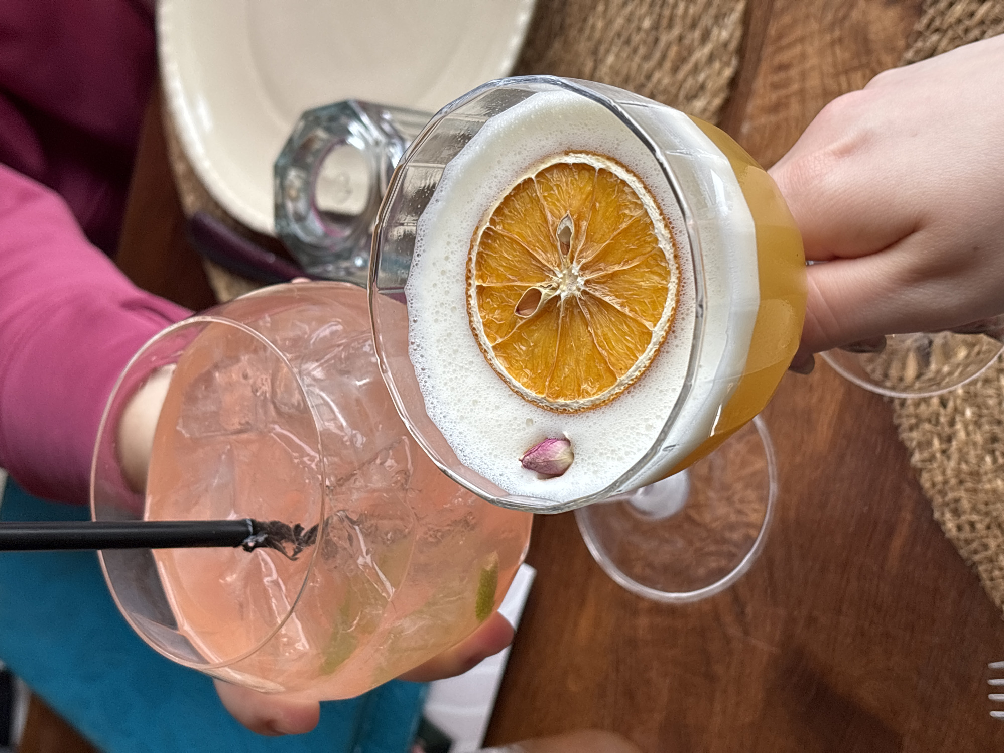 Two cocktails are presented. One cocktail is in a large, round glass filled with a pink liquid and ice, garnished with a lime wedge and featuring a black straw. The other cocktail is in a more geometrically shaped glass, topped with a white foam, an orange slice, and a small flower bud. The cocktails are held by two people, their hands visible. One person is wearing a pink shirt and blue pants, while the other person's clothing is not fully visible. A fork is visible in the bottom right corner of the image. The scene is set on a wooden table, with a woven table mat visible in the background. There's also a dish and a container visible in the background. There is no discernible evidence in the image to pinpoint the location as Varna, Bulgaria, or any other specific location.