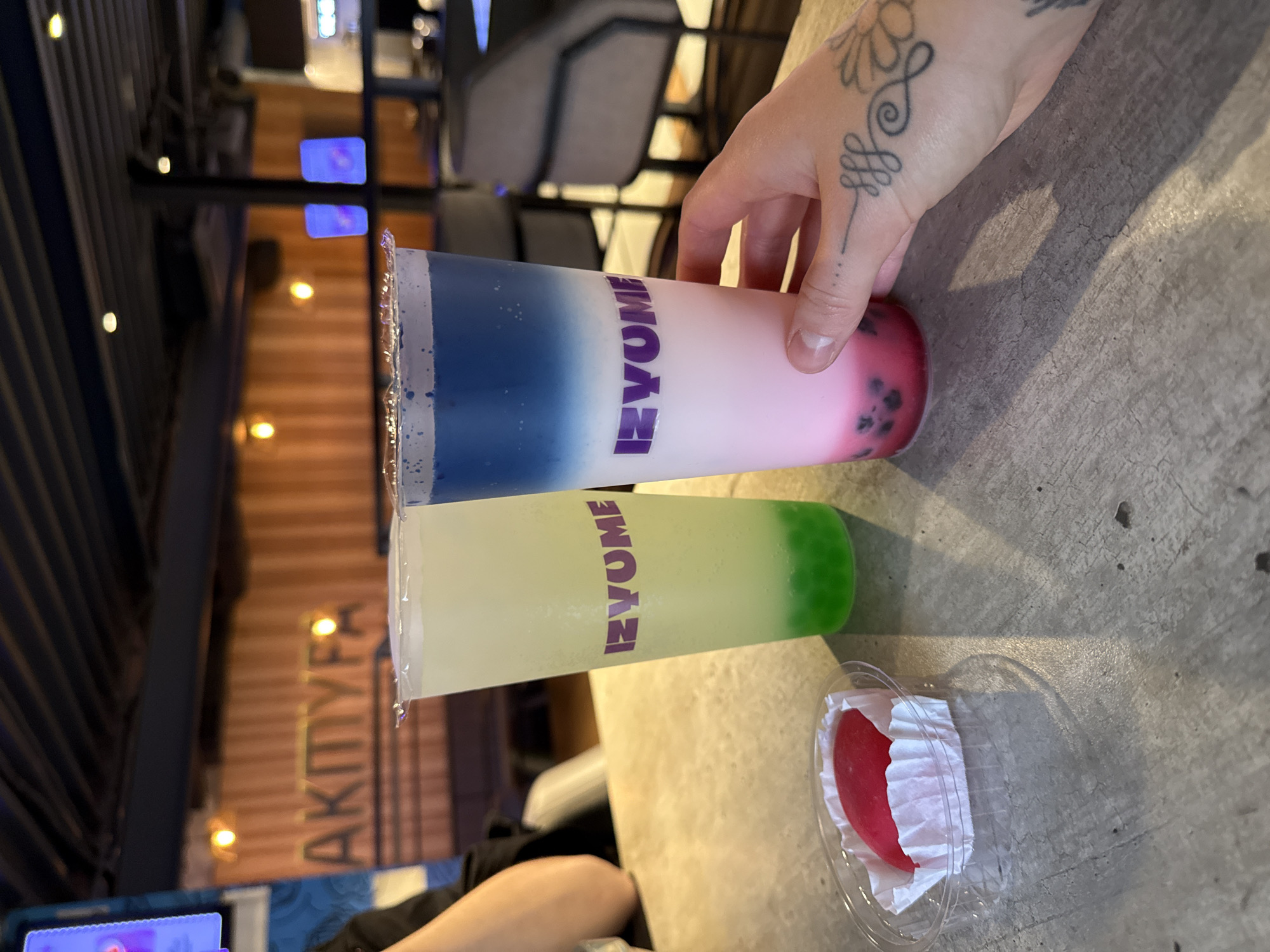 A table in a cafe in Rostov-on-Don, Russia, holds two transparent plastic cups of layered drinks, along with a clear plastic container holding a red object cushioned by white tissue. One cup is held by a hand with a tattoo, revealing the drink to be layered with blue at the top, transitioning to pink at the bottom, and contains dark-colored bobas. The second drink is green at the bottom, transitioning to clear liquid above. The cafe appears to be modern, with metal fixtures and a glimpse of an illuminated sign reading "АКПТУРА" visible in the background.