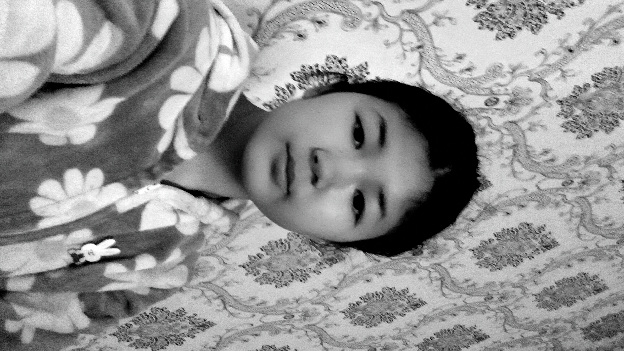 A black and white, head-and-shoulders selfie shows a young person with dark hair and fair skin in front of floral patterned wallpaper. The person is wearing a hooded jacket with a flower pattern and a small white rabbit emblem on the left side of the chest. The background wallpaper features a repeating ornate floral and vine motif in a lighter shade against a darker background. The scene appears to be indoors, likely a residential setting, with the lighting suggesting artificial light sources. No specific location within Uchkun, Kyrgyzstan, is discernible from the image itself.