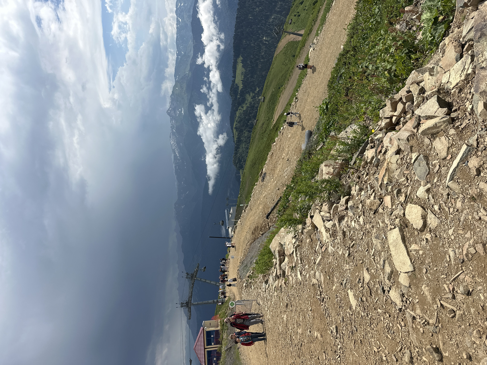 This image captures a scenic mountain landscape near Estosadok, Russia, likely at a ski resort during the warmer months. In the foreground, a dirt path, strewn with rocks and gravel, winds uphill. Several individuals are walking along this path. To the left, a red and yellow building, possibly a restaurant or information center, is visible, with ski lift infrastructure extending upwards behind it. Further into the background, rolling green hills give way to majestic, cloud-shrouded mountains with snow-capped peaks. The sky is a mix of dramatic dark clouds and brighter, sunlit areas, suggesting variable weather. The overall scene depicts a popular mountain destination with hiking trails and resort amenities.