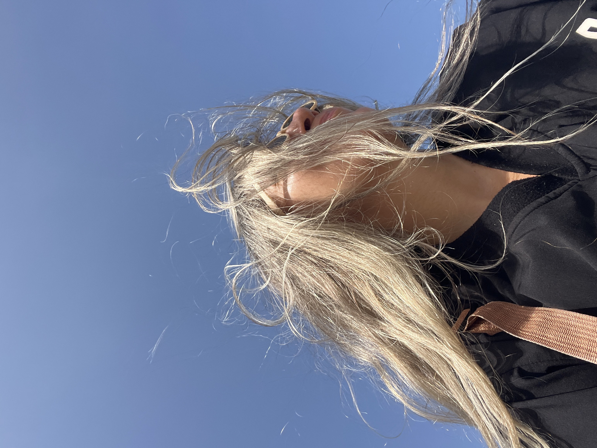 A medium shot captures a young woman with long, blonde hair and sunglasses against a clear blue sky. Her hair is blown back by the wind, partially obscuring her face, and she is wearing a black jacket with a tan strap across it. The shot is taken from a low angle, looking up at the woman. The background is an expansive, cloudless sky. No specific location in Matara, Sri Lanka is identifiable.