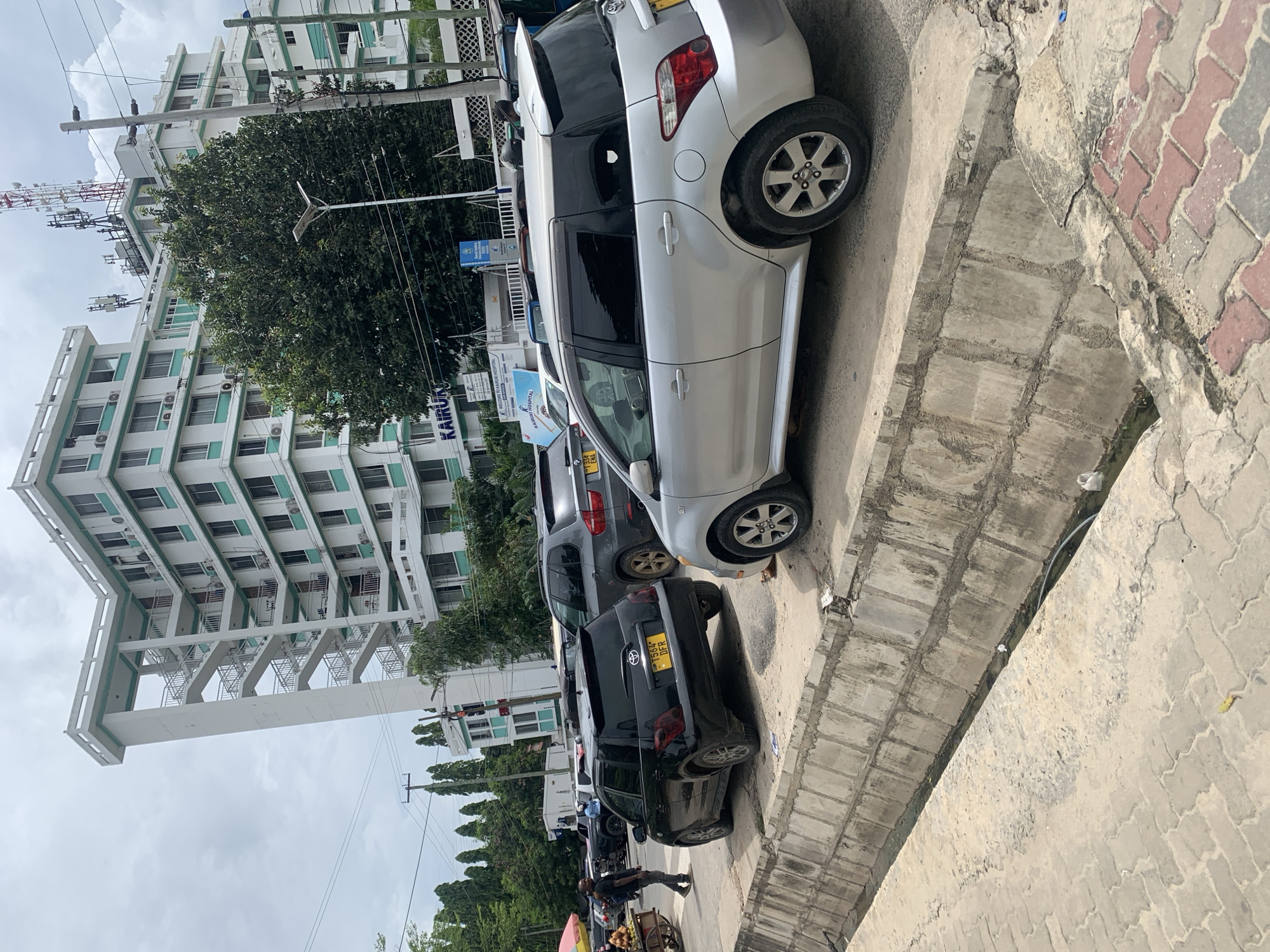 The image depicts a street scene in Dar es-Salaam, Tanzania. Parked along the right side of the street are several cars, including a silver Toyota, a gray SUV, and a black Toyota with a yellow license plate. A man is walking along the side of the road. Another man is walking on the sidewalk with a pushcart. On the left side of the street is a tall, white building with light green accents. A large tree is also visible, along with utility poles and overhead wires. The sky is overcast. The sidewalk is made of concrete pavers.