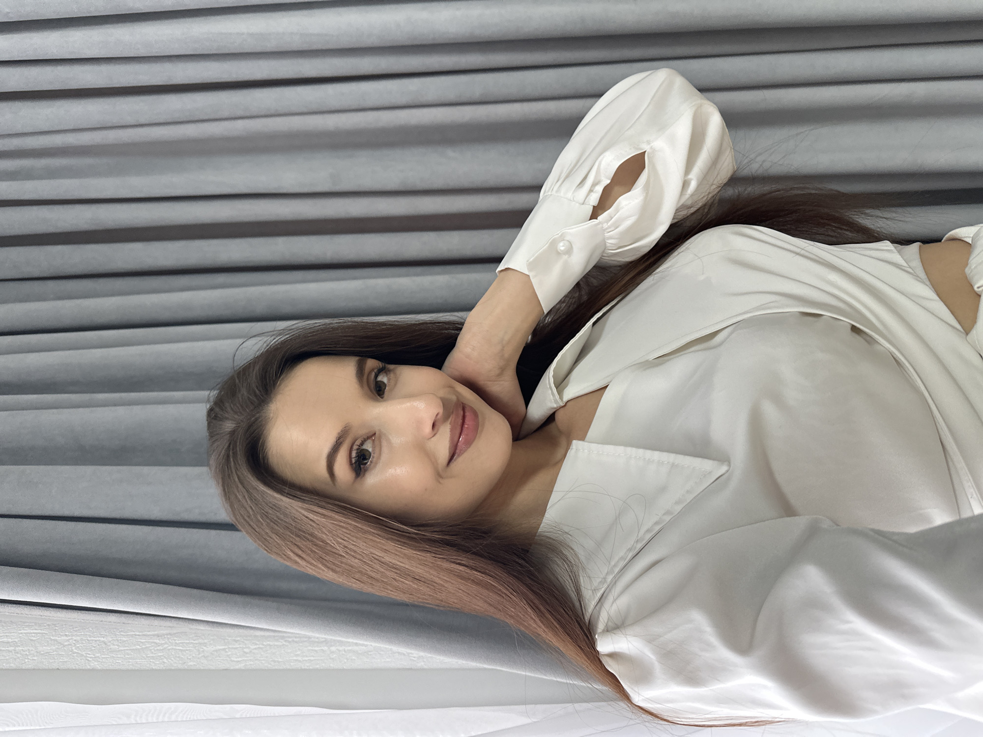 This indoor portrait features a young woman with long, flowing reddish-brown hair, looking directly at the camera with a gentle, engaging smile. She is seen from the chest up, positioned in a relaxed pose. Her right arm is raised, with her hand resting softly behind her neck, causing the sleeve of her top to gently billow.
She is wearing a white, satin-like long-sleeved shirt or blouse with a visible collar and cuffed sleeves, one of which features a small white button. Her hair cascades over her shoulders and chest. Her face is softly made up, highlighting her eyes and lips.
The background consists of uniformly pleated grey or silver fabric, likely curtains or blinds, creating horizontal textured lines. Below this, a plain white surface is visible. The lighting is soft and even, suggesting a bright indoor setting during the daytime. The woman's expression conveys a sense of calm and pleasantness. This scene is an indoor portrait captured in Yekaterinburg, Russia. There is no visible text in the image.