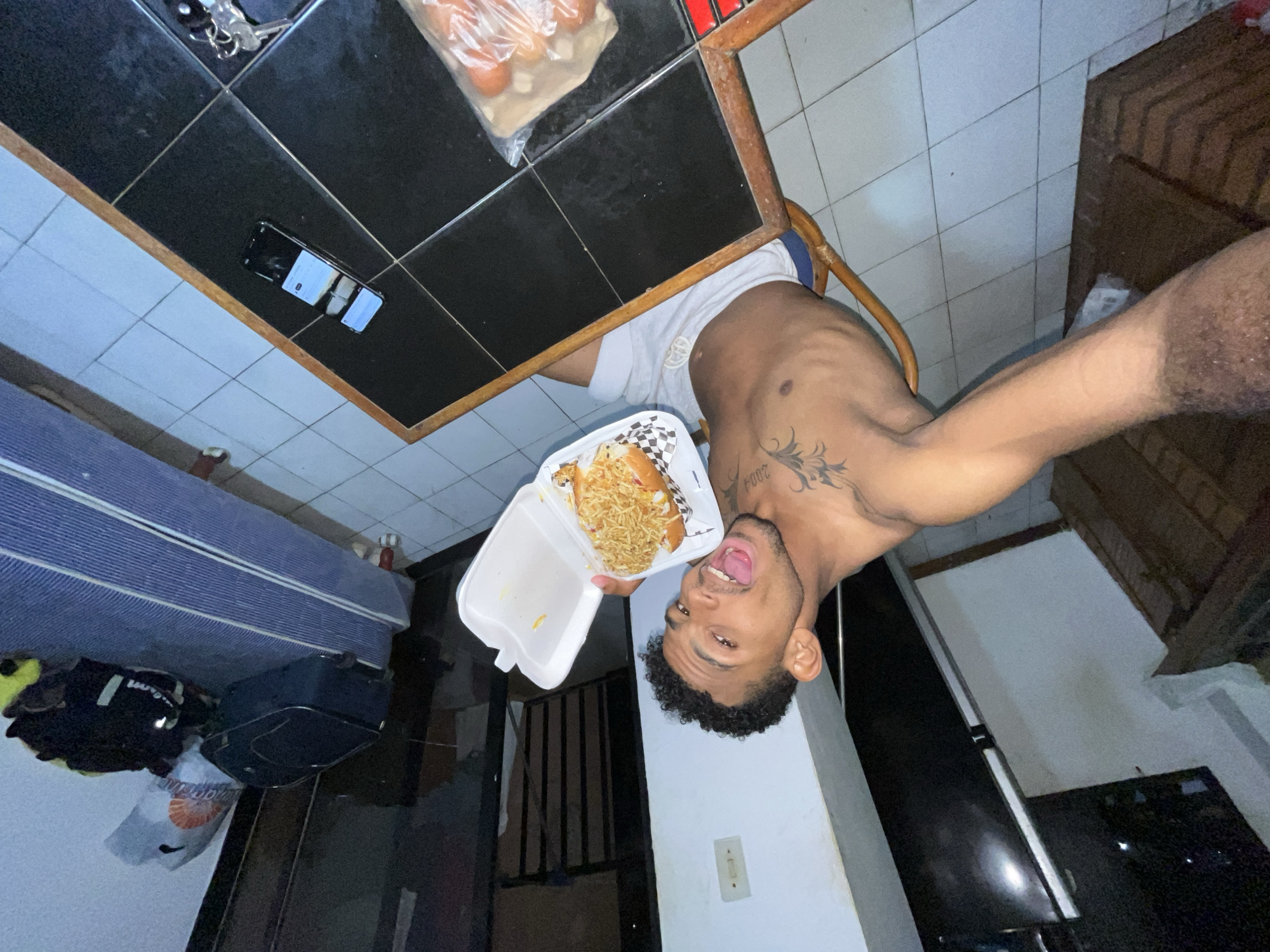 A young man playfully poses for a selfie indoors, holding a takeout meal. The image is oriented sideways, making the man appear upside down in the frame.
The man has dark, curly hair, a beard, and is shirtless, wearing white shorts. He has a large, dark tattoo on his left shoulder/upper arm, where the numbers "2004" are partially visible. His mouth is open with his tongue slightly out, and he looks directly at the camera, conveying a lighthearted and expressive mood.
He holds a white styrofoam container with a hot dog or similar sandwich, generously topped with shoestring fries or crispy noodles, and served on a checkered paper liner.
Behind him, on a table with a black tiled surface and a wooden border, are a smartphone with a cracked screen and a set of keys. A clear plastic bag containing eggs or similar round items is also on the table. The floor is tiled with light-colored square tiles.
In the background, a blue and white striped mattress is visible, along with a black duffel bag/suitcase, a yellow item of clothing, and a white plastic bag with an orange logo. A white wall features an electrical outlet, and a dark wooden object or doorway is visible. The indoor setting, with artificial lighting, suggests it might be evening or night. The scene depicts a casual moment in a residential space, consistent with the location context of Porlamar, Venezuela.