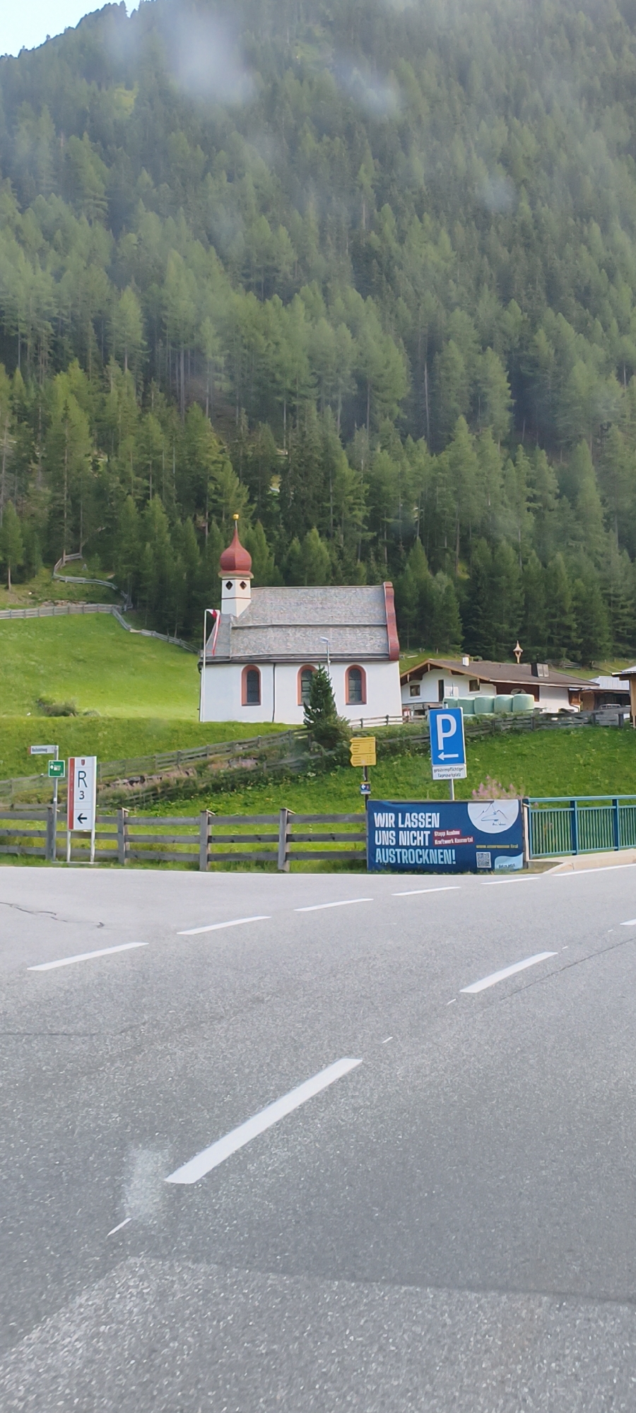 This is a medium shot, eye-level outdoor photograph taken in Sölden, Austria, featuring a small, white church with a red-tiled roof and a red spire. The church is situated on a grassy hillside, with a dense forest of green pine trees behind it, partially obscured by mist or clouds. A wooden fence runs along the lower edge of the hillside, separating it from a paved road in the foreground.
Several signs are visible. To the left of the church, a white sign with a green border and an arrow points to the left. Below it is a white sign with "R" and "3" in black. To the right of the church, a blue sign with a white "P" indicates parking. A horizontal banner with German text and an image of mountains is stretched across the fence on the right side of the road. There are no people visible in the image. The road surface is asphalt with white lane markings. In the background, there appear to be some buildings with similar red-tiled roofs. The overall atmosphere is somewhat overcast due to the mist or clouds.