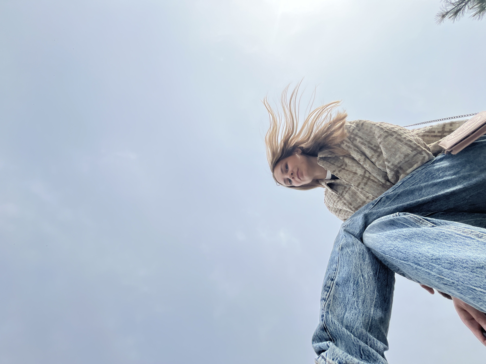 A low-angle shot shows a young woman in Chaikovsky, Russia. She wears light-wash jeans, a beige textured jacket, and a pink purse with a chain strap. Her blond hair is swept back, possibly by wind. The background is a pale sky.