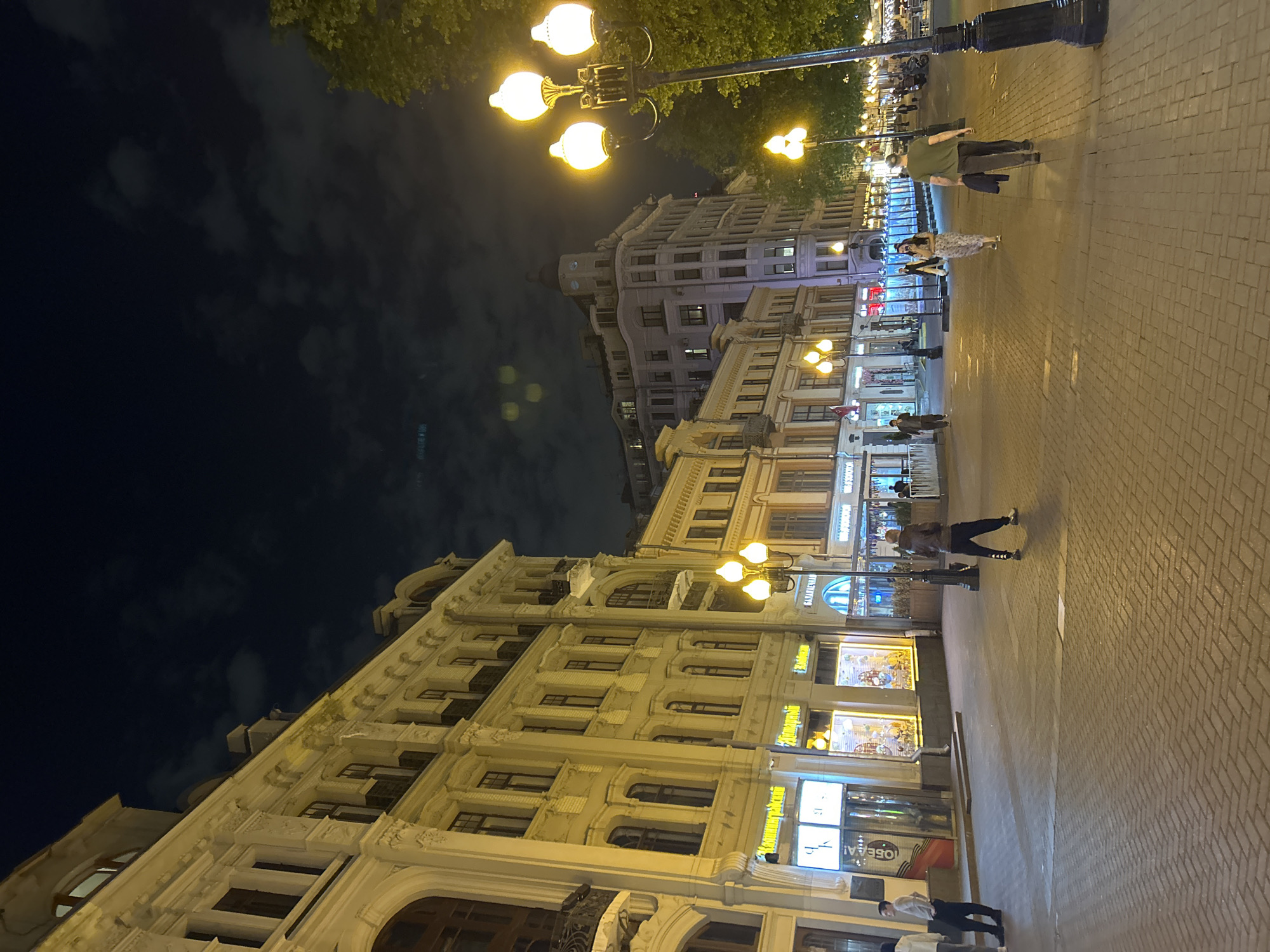 Captured at night, the image depicts a street scene in an urban setting, likely the city of Unknown, Unknown. The foreground is dominated by a pedestrian walkway paved with bricks. Several people are strolling along the walkway, while others are standing near shops. The buildings lining the street are illuminated with a mix of street lights and the glow from store displays. The architectural style of the buildings is diverse, with some structures displaying ornate details and historical facades. Above the buildings, the night sky is visible, punctuated by clouds. Street lamps equipped with multiple bulbs provide bright, warm light, enhancing the scene's overall ambiance.