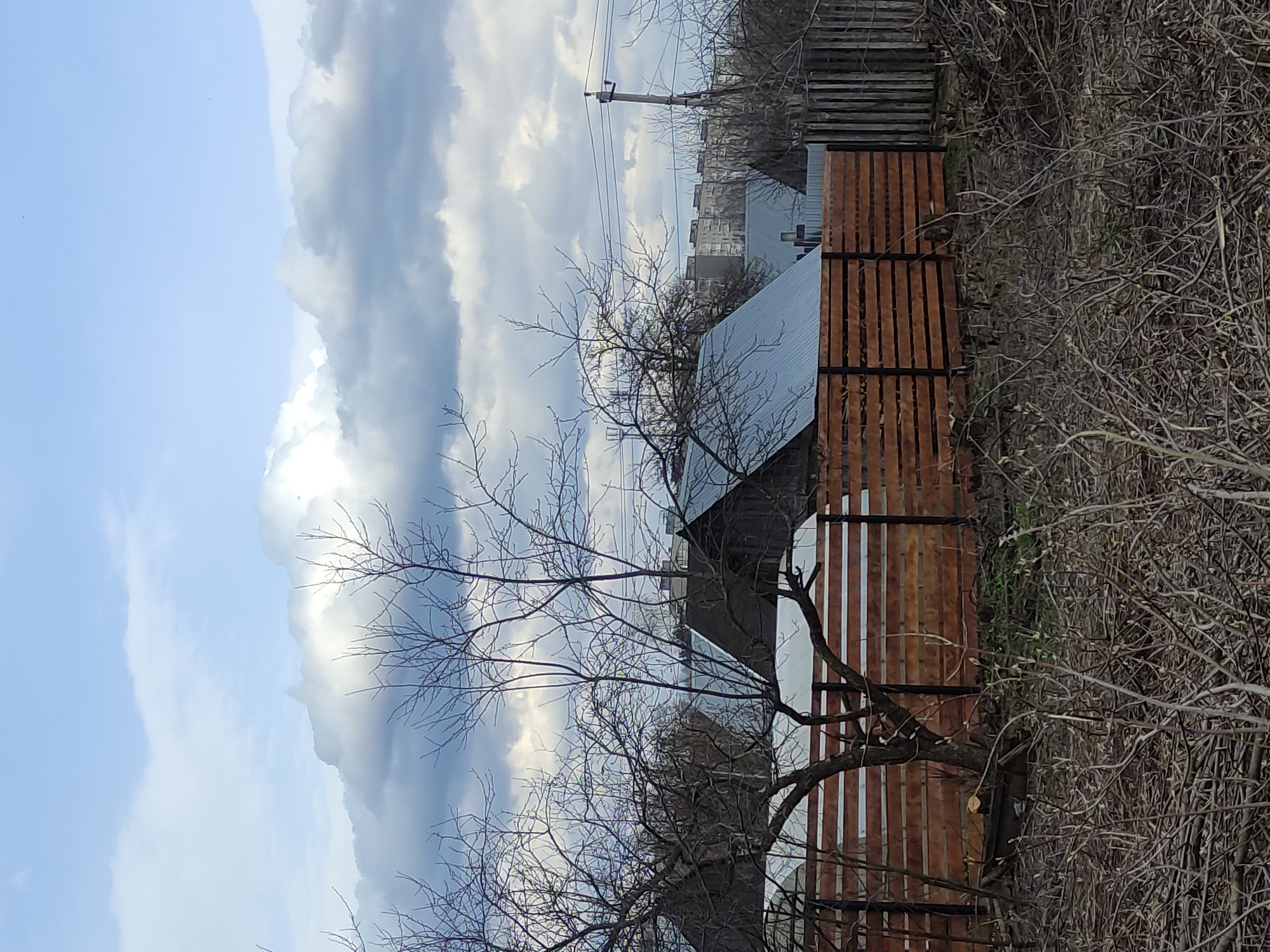 The image depicts a view of a residential area in the city of Unknown, Unknown. A brown wooden fence stands on the right side of the frame, partially obscured by bare branches of a tree. Behind the fence, a building with a metal roof is visible. In the background, taller buildings can be seen under a sky filled with clouds. A utility pole with wires is present in the distance. The scene is captured during what appears to be a season with leafless trees, suggesting autumn or winter.