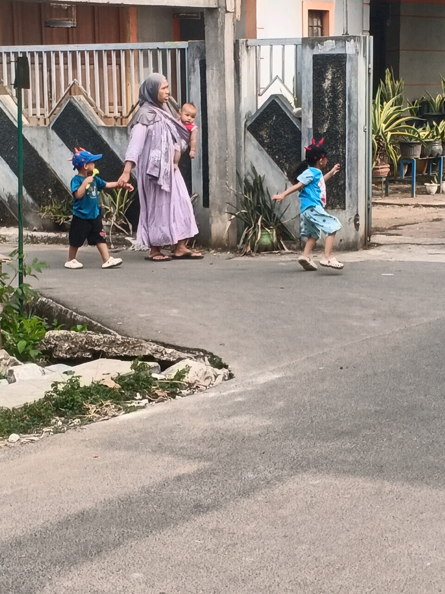 The image depicts a street scene in Kadu, Indonesia, on a bright day. A woman wearing a lilac hijab and a flowing dress walks to the left, carrying a baby in a sling on her back. Beside her, a young boy in a blue shirt and black shorts, wearing a blue cap with red accents, holds her hand. He has a light-colored pacifier in his mouth. To the right of the woman, another child, a girl in a blue shirt and patterned shorts with red devil horns on her head, runs ahead. She is wearing white sandals. The children are on the asphalt road. Behind them, a metal gate with patterned panels is visible, leading to a property with a building. Green plants in pots are situated to the right of the gate. The foreground of the image shows the edge of the road with some overgrown greenery and debris. The overall scene suggests a casual outing or walk in a residential area.