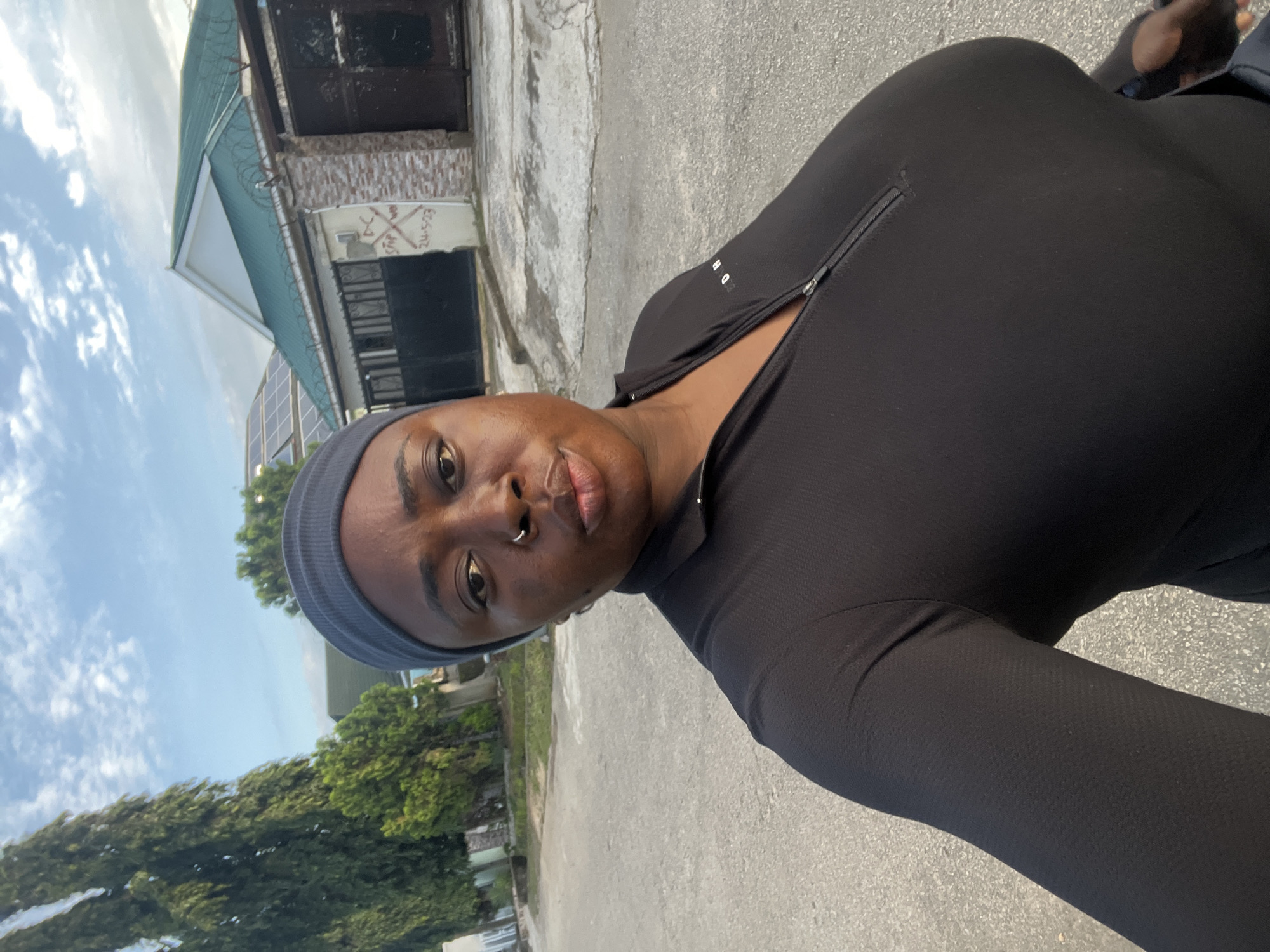 A woman is captured in a selfie outdoors, with a city street and residential buildings in the background. The woman is wearing a black long-sleeved top with a zippered neckline and a grey headscarf. She has a nose ring and appears to be looking at the camera. The environment suggests a residential area in Abuja, Nigeria, with paved roads, trees, and houses with green roofs and security features like barbed wire. The sky is partly cloudy, indicating daylight.