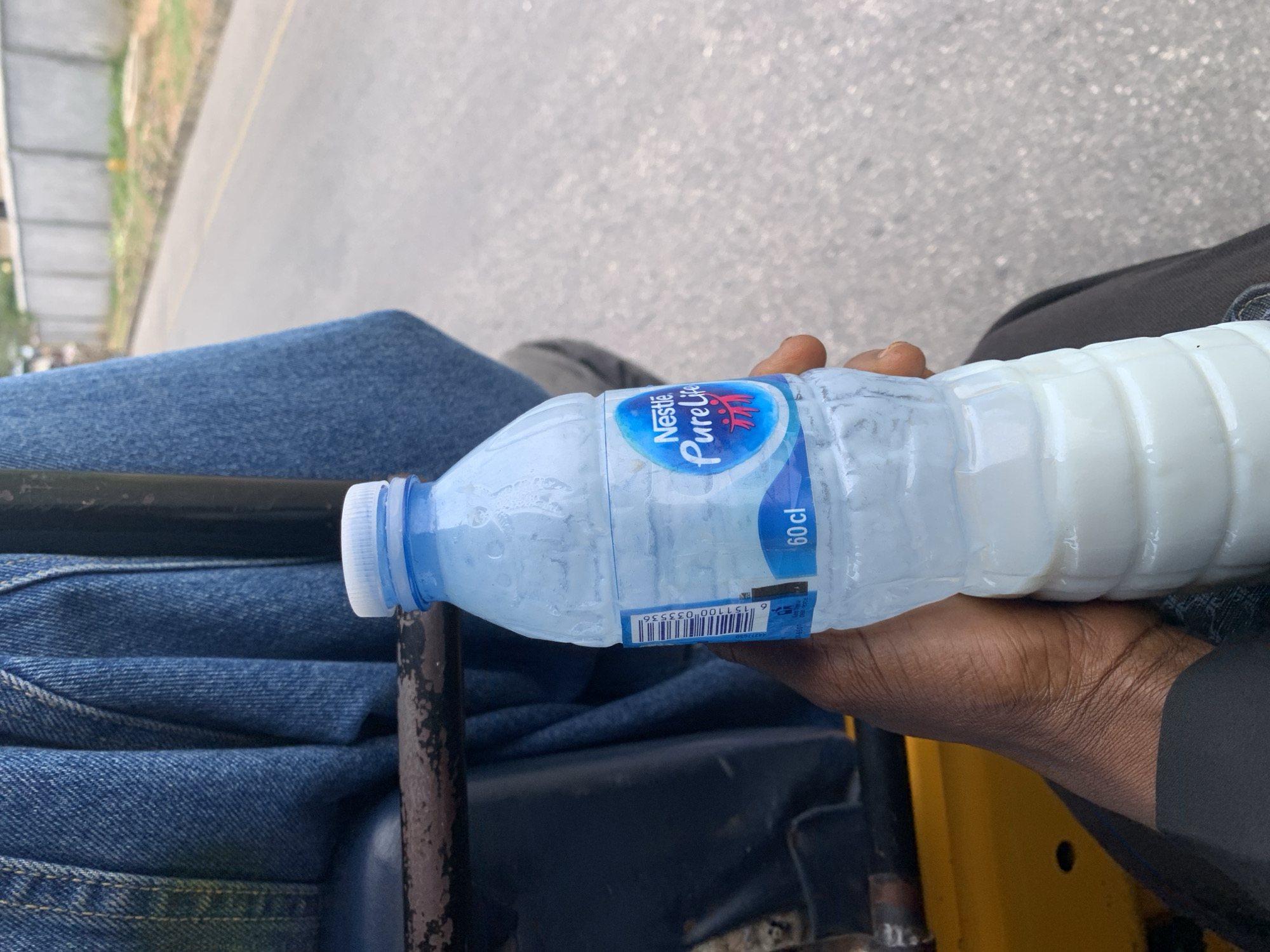 A first-person, close-up perspective shows a person seated in what appears to be a vehicle, likely a motorcycle or tricycle, in Lokoja, Nigeria. The person is holding a partially filled, clear plastic bottle of Nestle Pure Life drinking water. The bottle is approximately 60 cl in size, with a blue label. The liquid inside the bottle is white. The person wears blue jeans and a dark-colored jacket. The vehicle's handlebars and part of the vehicle's frame are visible. The background shows an asphalt road and some greenery, suggesting an urban or semi-urban setting.