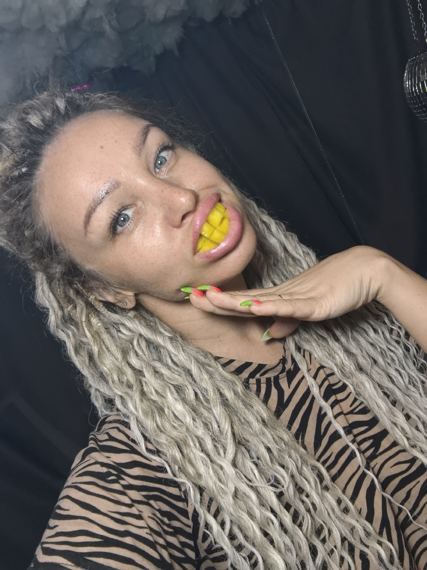 A woman with light-colored dreadlocks and blue eyes, wearing a tan and black zebra-striped shirt, poses for a selfie. She is holding her hand up to her face with brightly colored nails. Yellow objects are placed in her mouth to resemble teeth. The background is dark, and a disco ball hangs to the right. There is no evidence to support the location being Noviy, Russia.