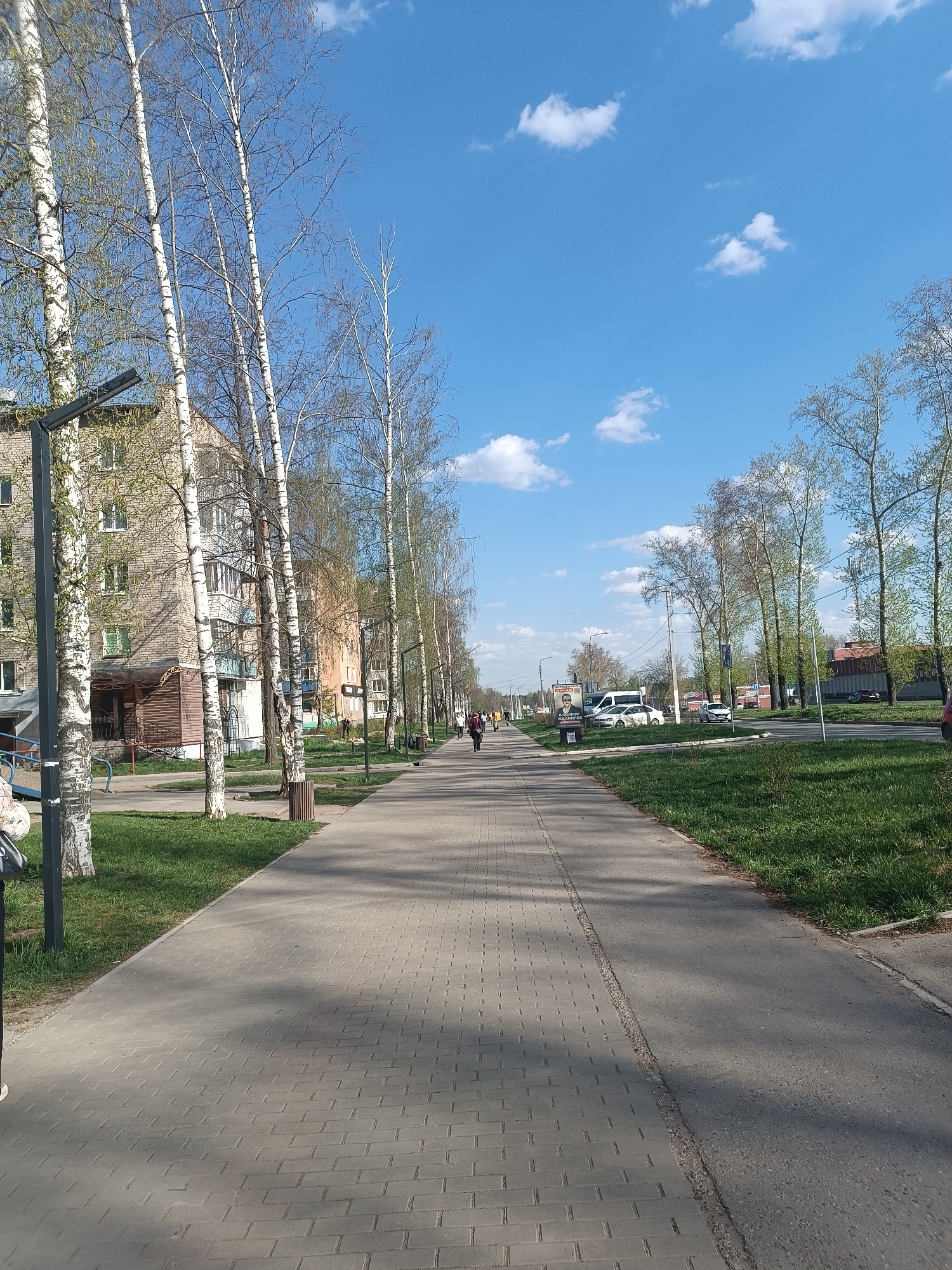 A straight paved pedestrian walkway extends into the distance, flanked by grassy areas and trees. The trees, mostly birch, line the walkway, their canopies just beginning to leaf out. Buildings are visible to the left, while cars and other structures can be seen on the right side of the path. Several people can be seen walking along the walkway. The sky is blue with scattered clouds. The scene is in Chaikovsky, Russia.