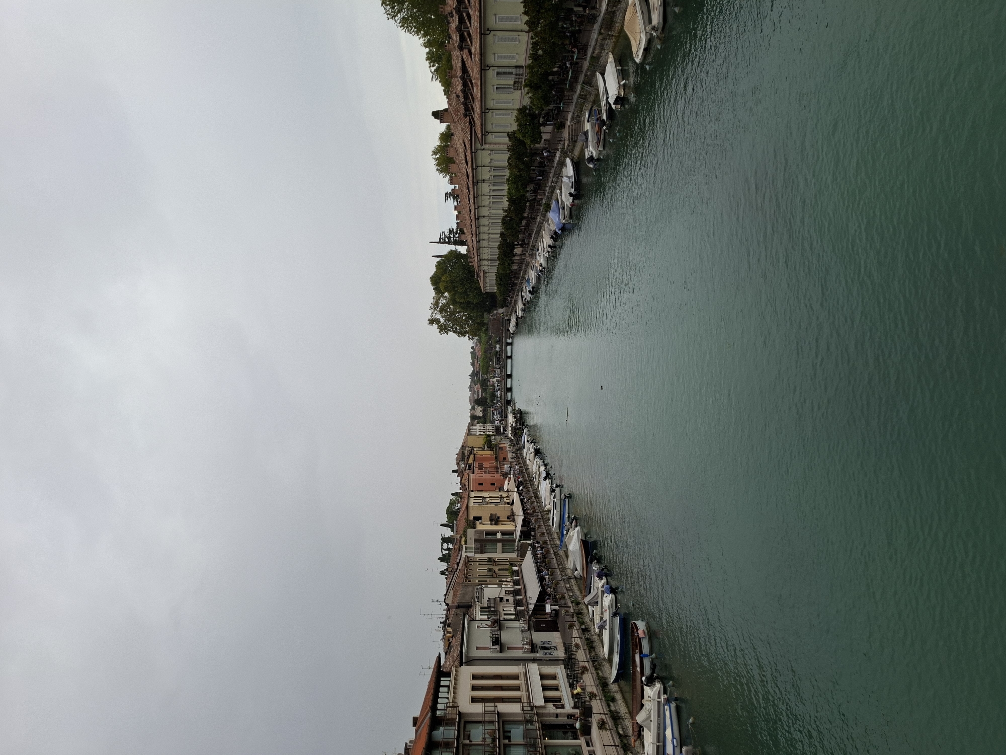 The image depicts a wide canal in Peschiera del Garda, Italy, lined with buildings and docked boats on both sides. The canal runs from the foreground into the distance, where a bridge can be seen. The sky is overcast with a grey, cloudy appearance.
On the left bank of the canal, there are multi-story buildings with balconies and windows. Numerous boats of various sizes are moored along the embankment. Some of these boats are covered with tarps.
On the right bank, a row of long, uniform buildings with terracotta tiled roofs is visible. These buildings appear to be residential or commercial establishments. Similar to the left bank, boats are docked along the edge of the canal in front of these buildings. Trees are planted along the walkway in front of the buildings on the right.
The water in the canal is a deep turquoise-green color and appears calm, with slight ripples visible. There are a few dark objects that could be birds or ducks swimming in the water. No people are clearly visible in the image, although there are indications of outdoor seating areas on the banks suggesting the presence of cafes or restaurants. The overall scene suggests a tranquil, albeit somewhat overcast, day in a waterfront town.