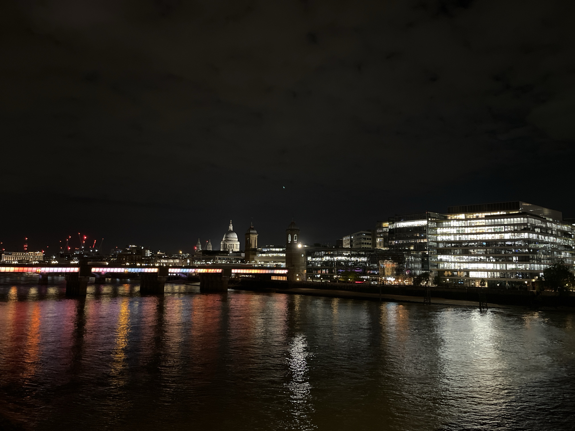 This is a nighttime photograph of the River Thames in London, United Kingdom, viewed from the South Bank looking north. The foreground features the dark, undulating surface of the river, reflecting numerous artificial lights from the city. In the midground, a multi-piered bridge, likely Southwark Bridge, spans the river, brightly illuminated along its length with a spectrum of colors including red, orange, yellow, pink, blue, and purple, all mirrored in the water below.
On the far bank (north bank), the cityscape is brightly lit. To the left of the bridge, the prominent dome of St. Paul's Cathedral is visible against the night sky, flanked by other illuminated historical buildings and distant red-lit construction cranes. To the immediate right of the bridge, a clock tower and another historical-looking tower stand out, their lights reflecting on the water. Further to the right, a large, modern, multi-story office building with numerous brightly lit windows dominates the right side of the frame, its light also reflecting in the river. The sky above is dark and cloudy, with discernible cloud patterns, and a single faint green light, possibly an aircraft, is visible in the upper mid-portion of the sky. No human figures are distinctly visible.