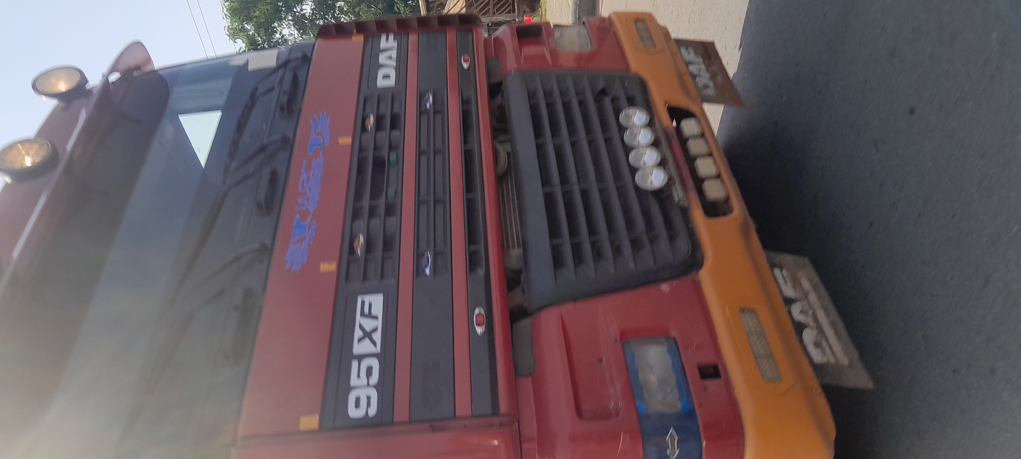 A red DAF 95XF truck is seen from the front, with its grille and orange bumper prominently displayed. The truck appears to be in motion, suggesting it is on a road. The background shows a slightly out-of-focus environment with trees and a structure that could be part of a building or a roadside stall. The lighting suggests it is daytime. The scene is likely within the city of Jalingo, Nigeria, given the context provided.