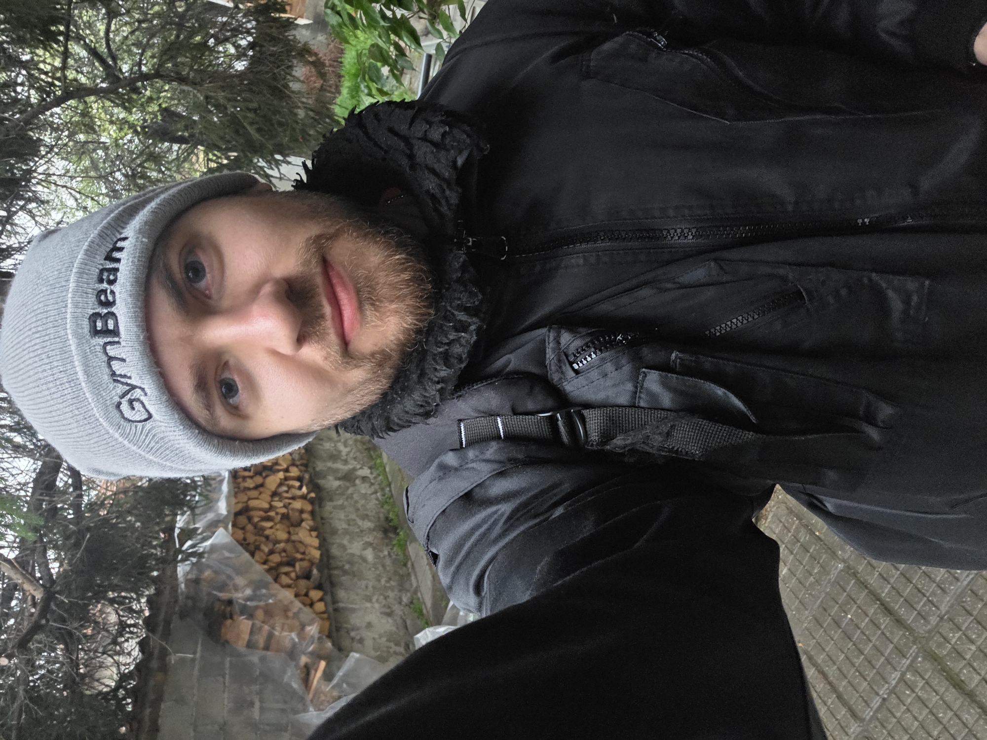 A medium shot captures a man with light skin, a beard, and light-colored eyes, wearing a grey beanie with "GymBeam" printed on it. He is also wearing a black jacket over a black garment with a strap. Behind him is a wall partially covered with plastic sheeting, and stacks of split firewood are visible. There are trees and greenery visible in the background. The location cannot be confirmed as Yagodovo, Bulgaria, based on the visual information.
