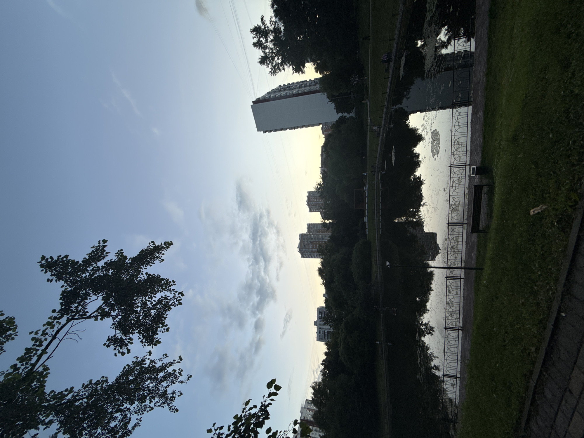 The image depicts a twilight scene at a park in Moscow, Russia, featuring a serene pond surrounded by lush greenery and residential buildings. The sky is a soft blend of blue and grey, with wispy clouds indicating the end of the day. Silhouetted trees frame the upper left portion of the image, their leaves gently rustling in the breeze.
In the background, several apartment buildings rise above the tree line, their modern architecture contrasting with the natural elements of the park. One tall, white building stands out on the right, its reflection visible in the still waters of the pond. The pond itself is dark and reflective, with scattered lily pads and the mirror-like image of the trees and buildings.
A black metal fence runs along the edge of the pond, separating it from a paved walkway and a grassy area. A park bench is visible on the grass, and a few individuals can be seen strolling along the walkway, their figures small and indistinct in the fading light. The overall atmosphere is peaceful and tranquil, characteristic of a city park at dusk.