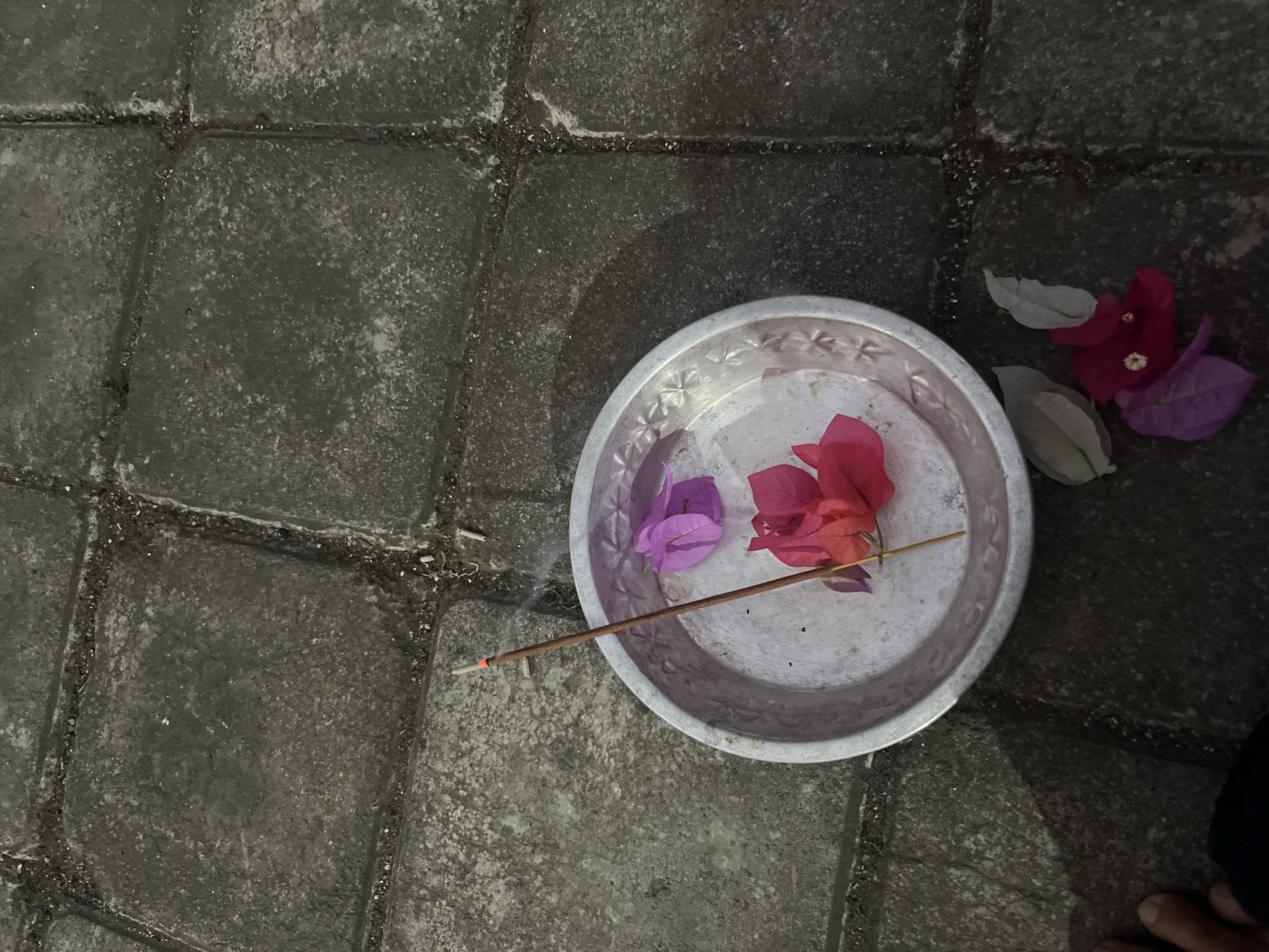 An overhead shot shows a small, round, metallic dish containing two bright pink bougainvillea petals and one purple bougainvillea petal. A single incense stick, lit and emitting a thin wisp of smoke, is placed in the dish. The dish is positioned on a stone patio with rectangular pavers. Several more bougainvillea petals, some pink and some light gray, are scattered on the patio stones to the right of the dish. A person's bare foot is visible in the lower left corner of the frame. The scene is likely in the city of Mendoyo, Indonesia, based on the presence of bougainvillea and the common practice of offering incense and flowers.