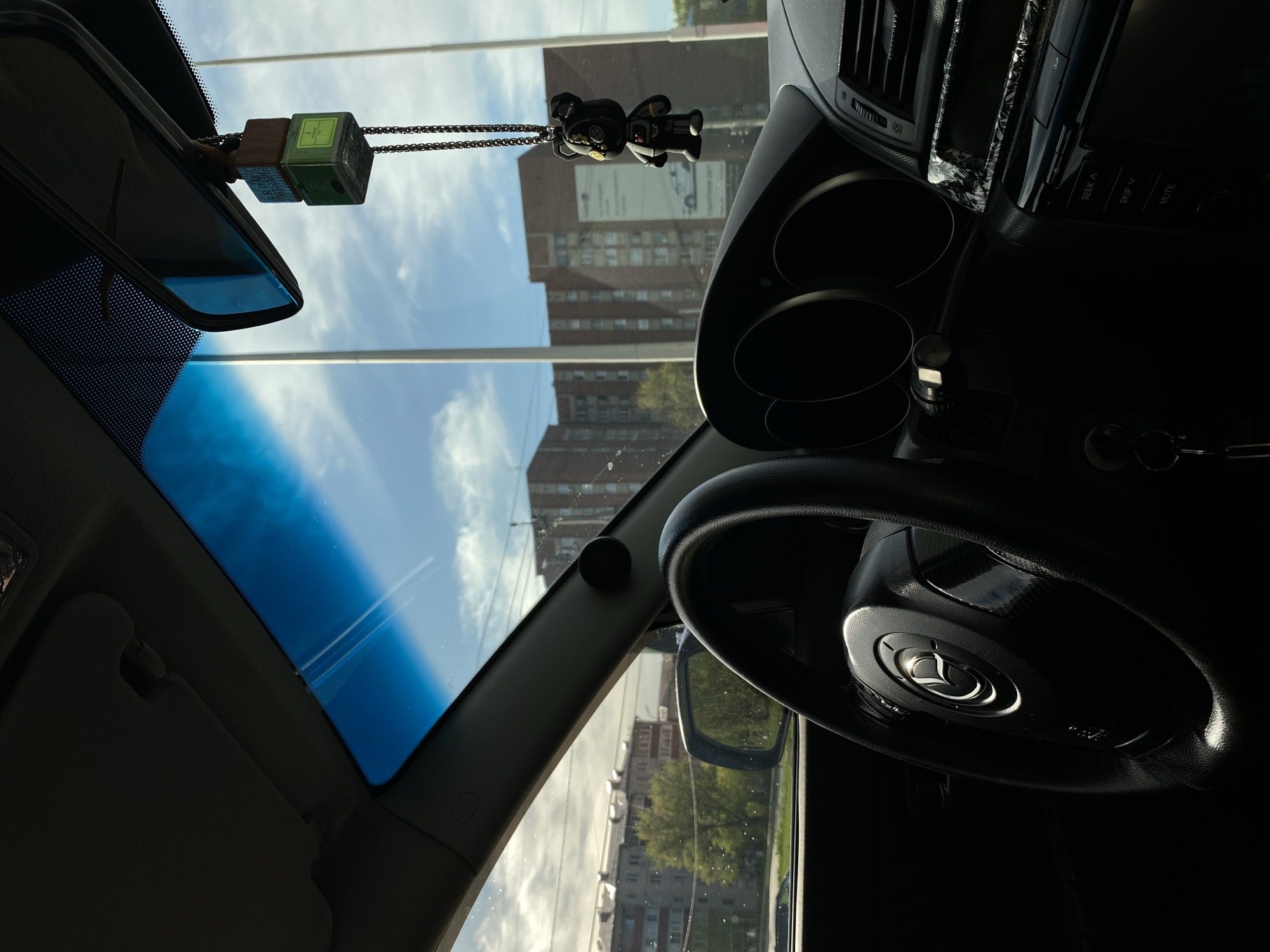 The image depicts the interior of a car, specifically the dashboard, steering wheel, rearview mirror, and windshield. Hanging from the rearview mirror are two items: a small green air freshener cube and a black bear-shaped charm. Outside the windshield, buildings are visible, indicating an urban environment. Based on the architectural style, the location appears to be in Votkinsk, Russia. The car is likely parked, and there are no visible people in the frame.
