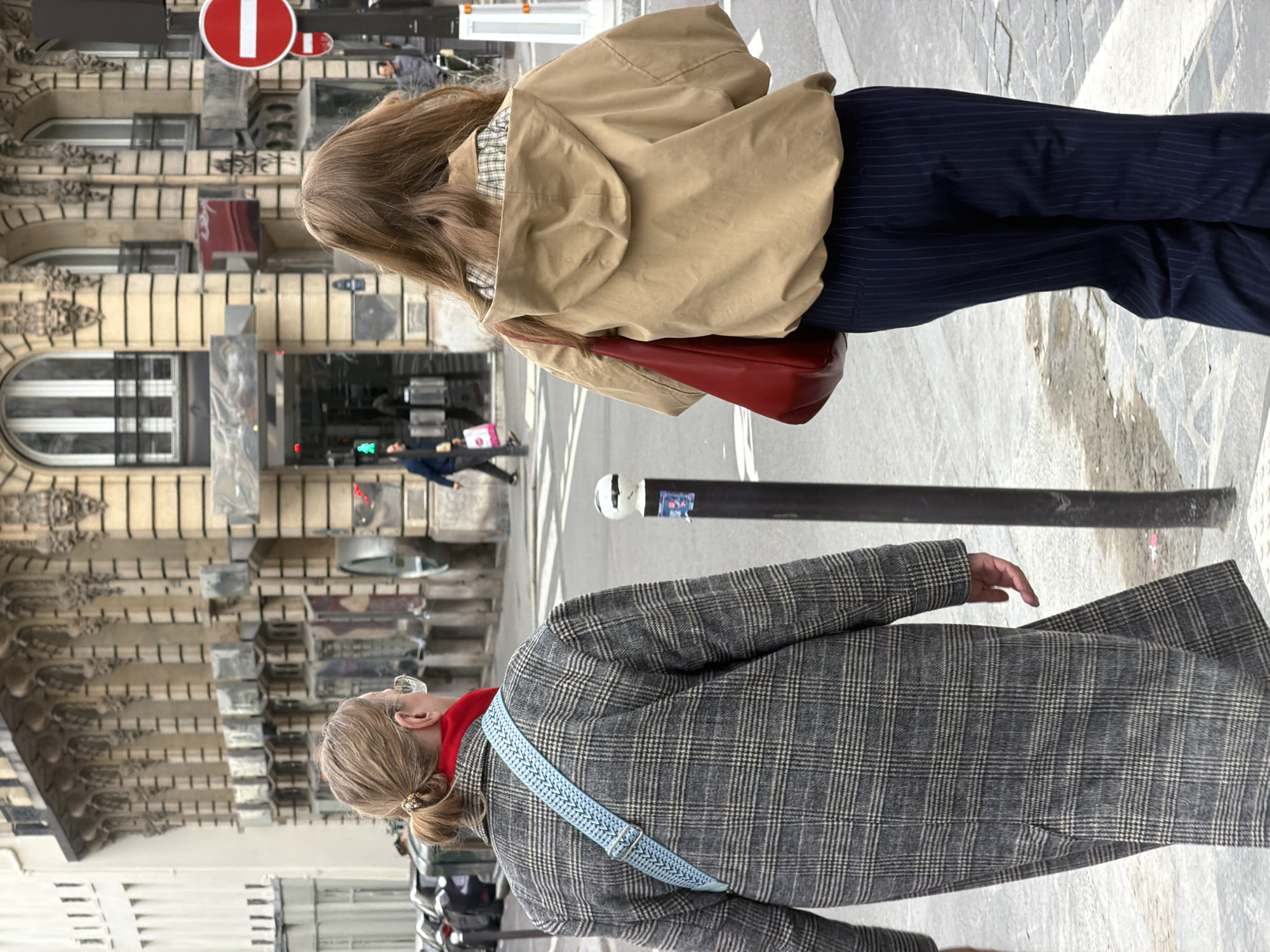 Two women are standing on a sidewalk near a street in Paris, France. The woman in the foreground wears a gray and white plaid coat, a red turtleneck sweater, and a blue and white strap bag. The woman in the background wears a tan jacket, dark blue pinstripe pants, and a red handbag. A street sign with a red circle and a white horizontal line is visible in the background. Pedestrians are crossing the street in the background. The architecture of Parisian buildings is visible in the background. A black pole with a white spherical cap is near the center of the image.