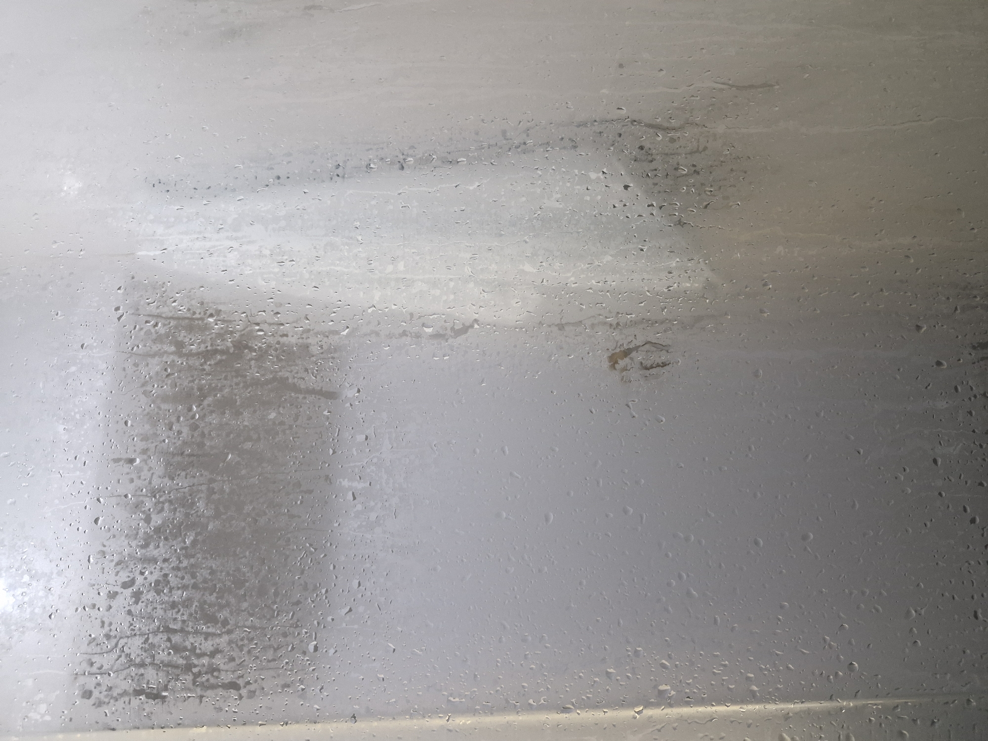 The image shows a close-up view of a wet, foggy glass surface, likely a shower door or window. Small water droplets are distributed across the glass, with some areas appearing more fogged than others. In the upper left portion of the image, a faint, blurred outline of what might be vertical blinds or a textured surface is visible through the condensation. A small, indistinct brown object, possibly a smudge or debris, is present in the central right area of the glass. The lighting appears diffused, creating a soft, hazy effect. The scene is entirely indoors, with no visible elements to suggest a specific location in Bulan-Segettu, Kyrgyzstan.