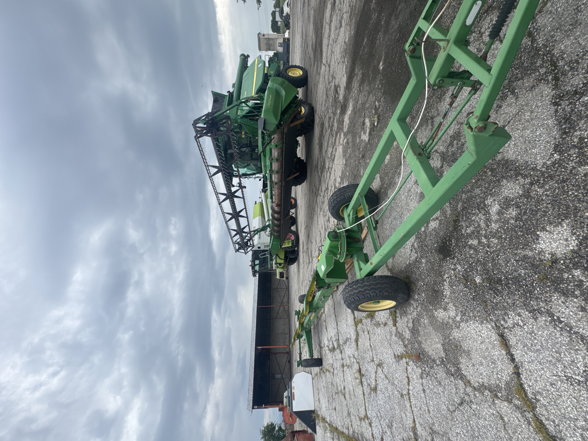 The image shows a green John Deere combine harvester parked on a paved surface in what appears to be an outdoor storage or maintenance area. Adjacent to the combine is a green equipment trailer with tires. In the background is a large metal shed or storage building and additional vehicles, possibly other farm equipment. The sky is overcast. The location is suggested to be Sarnevo, Bulgaria. No people are visible.