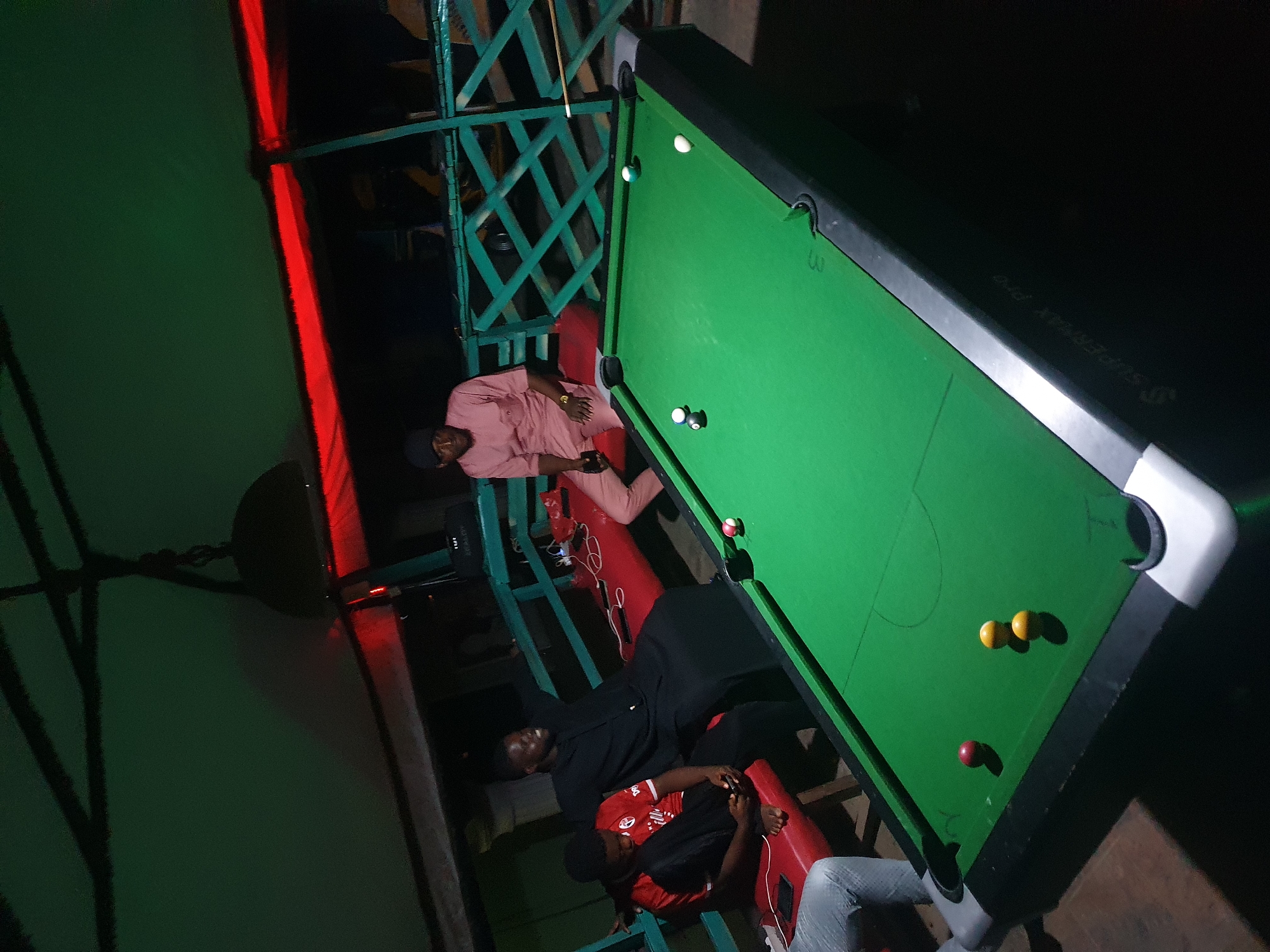 The image depicts a nighttime scene likely indoors at a pool hall, in Lokoja, Nigeria. A pool table dominates the view, with several billiard balls scattered on its green felt surface. Three men are seated on red cushioned chairs next to the pool table. One man is wearing a pink outfit, another is wearing a black outfit, and a third is wearing a red and black outfit. A portion of a fourth person can be seen. The lighting is dim, with some sections illuminated by a red light source and others shrouded in darkness. There are wooden cross-lattices near the pool table. The pool table has white corner pockets and the logo "SUPERMAX Pro".