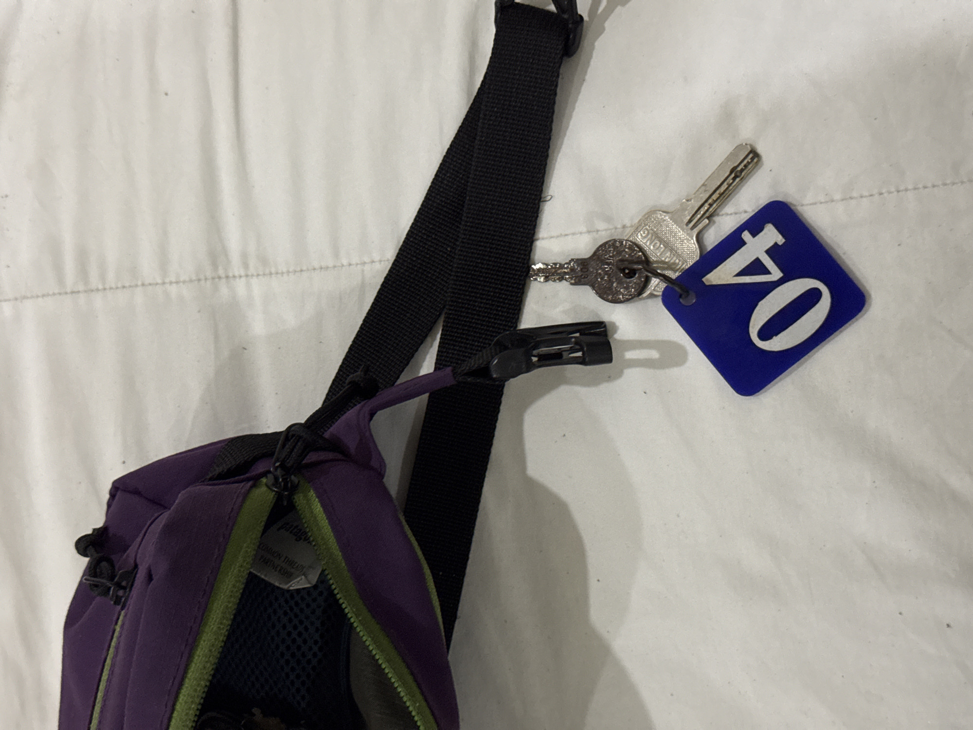 The image shows a purple and green bag with a black strap hanging on a white sheet. Attached to the strap is a set of keys with a blue keychain with the number "04" on it. The scene is indoors. There is no evidence to indicate the location is Đà Nẵng, Vietnam.