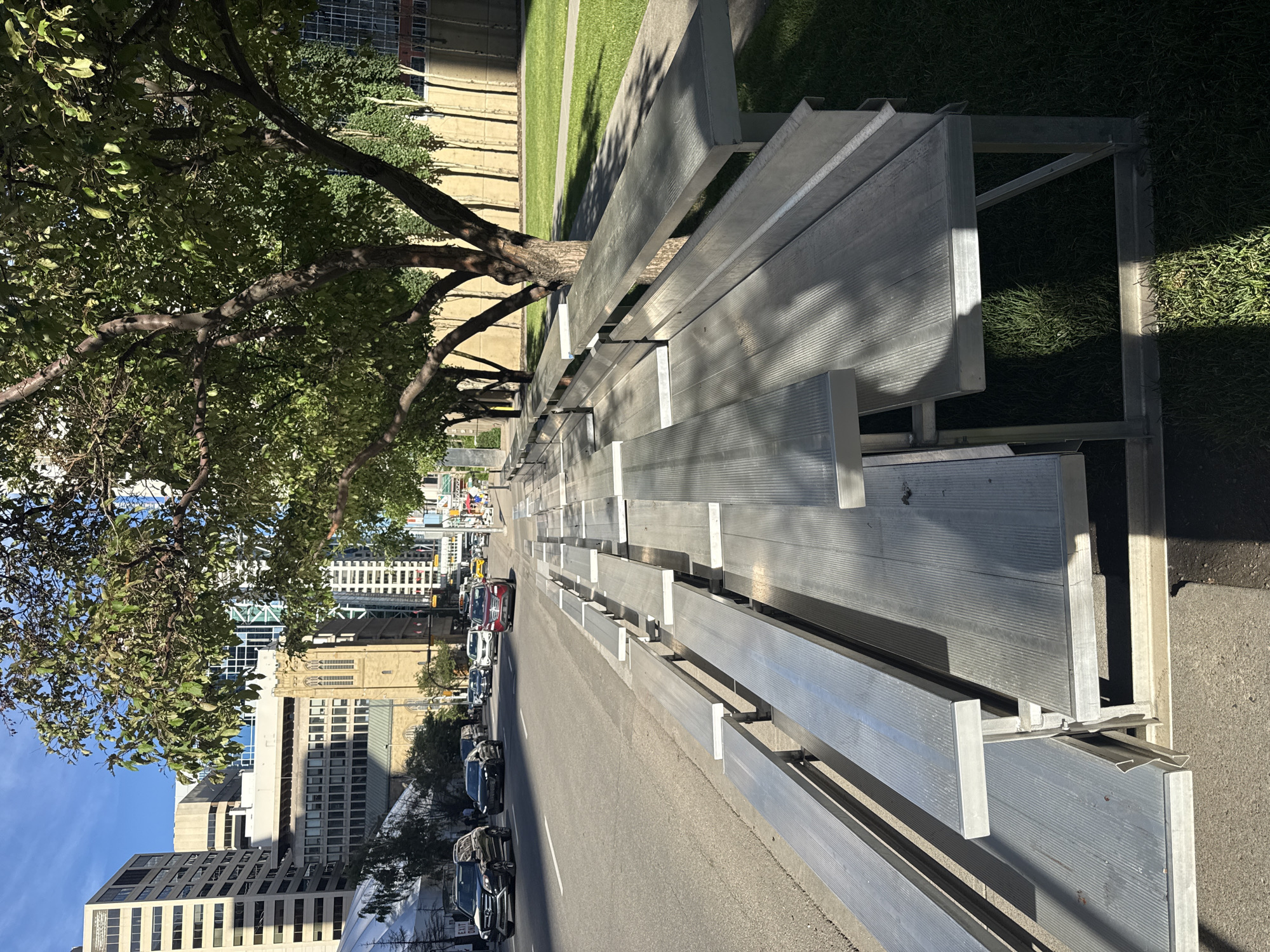 The image depicts a sunny day in Calgary, Canada, with a row of empty aluminum bleachers on the right side of a city street. The bleachers are positioned alongside a sidewalk, which separates them from a grassy area and a concrete wall. Beyond the grassy area, there are trees with lush green leaves. The street is moderately busy with cars driving on it, and buildings of various heights and architectural styles, including office buildings and a church with a prominent tower, are visible in the background. The lighting suggests it is likely late morning or early afternoon due to the shadows cast by the trees and bleachers. There are no people visible in the immediate foreground or on the bleachers themselves.