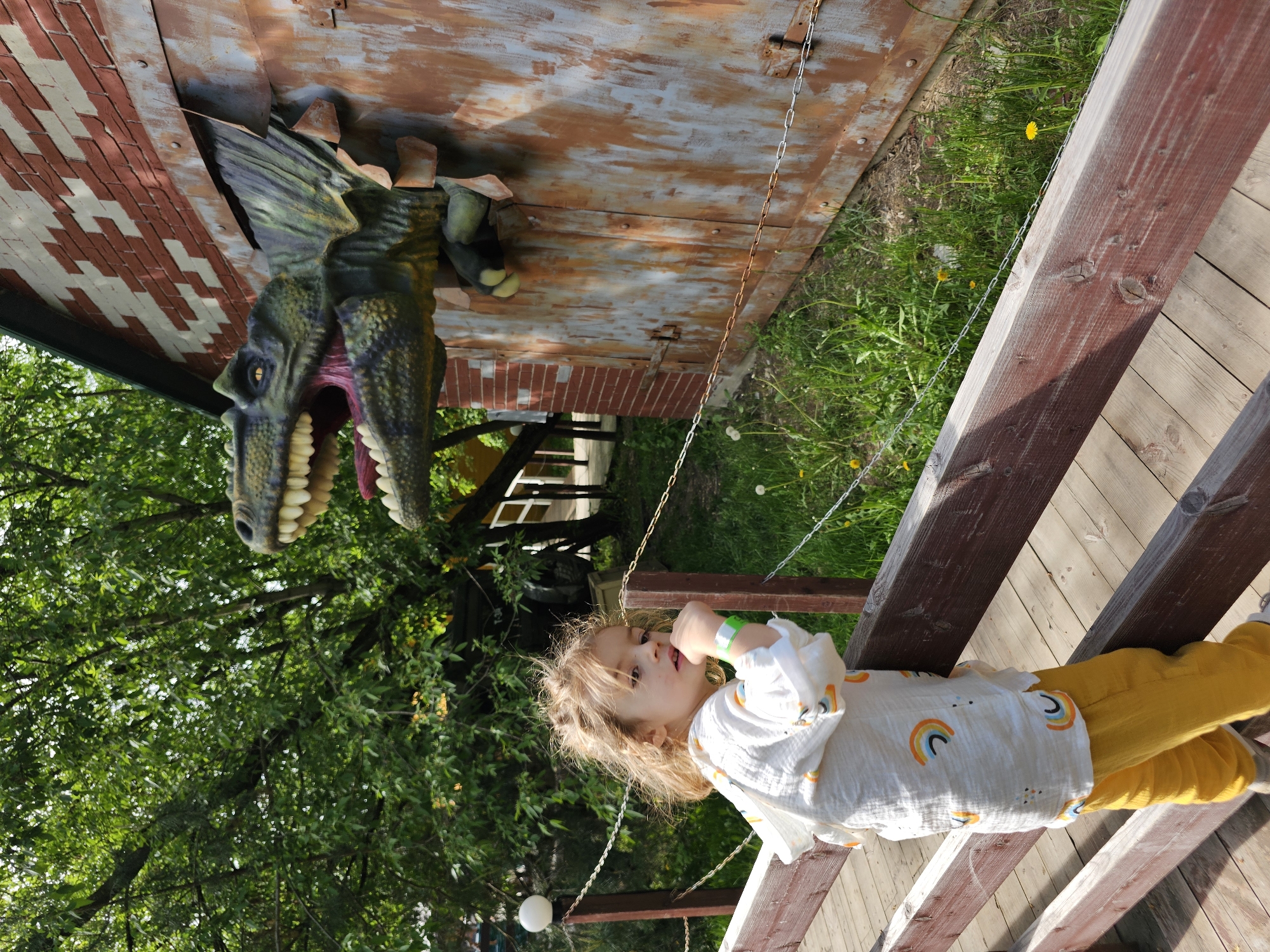 A child is seen on a wooden structure in what appears to be a park, as evidenced by the presence of grass and trees. The location is Khimki, Russia. A large dinosaur head protrudes from a building behind the child. The child is wearing a white shirt with rainbow patterns and yellow pants. The building has a brick and rusted metal exterior. There is a chain hanging down near the child and some white spherical lamps in the background, possibly indicating a decorative lighting system. The wooden structure has a reddish-brown hue.