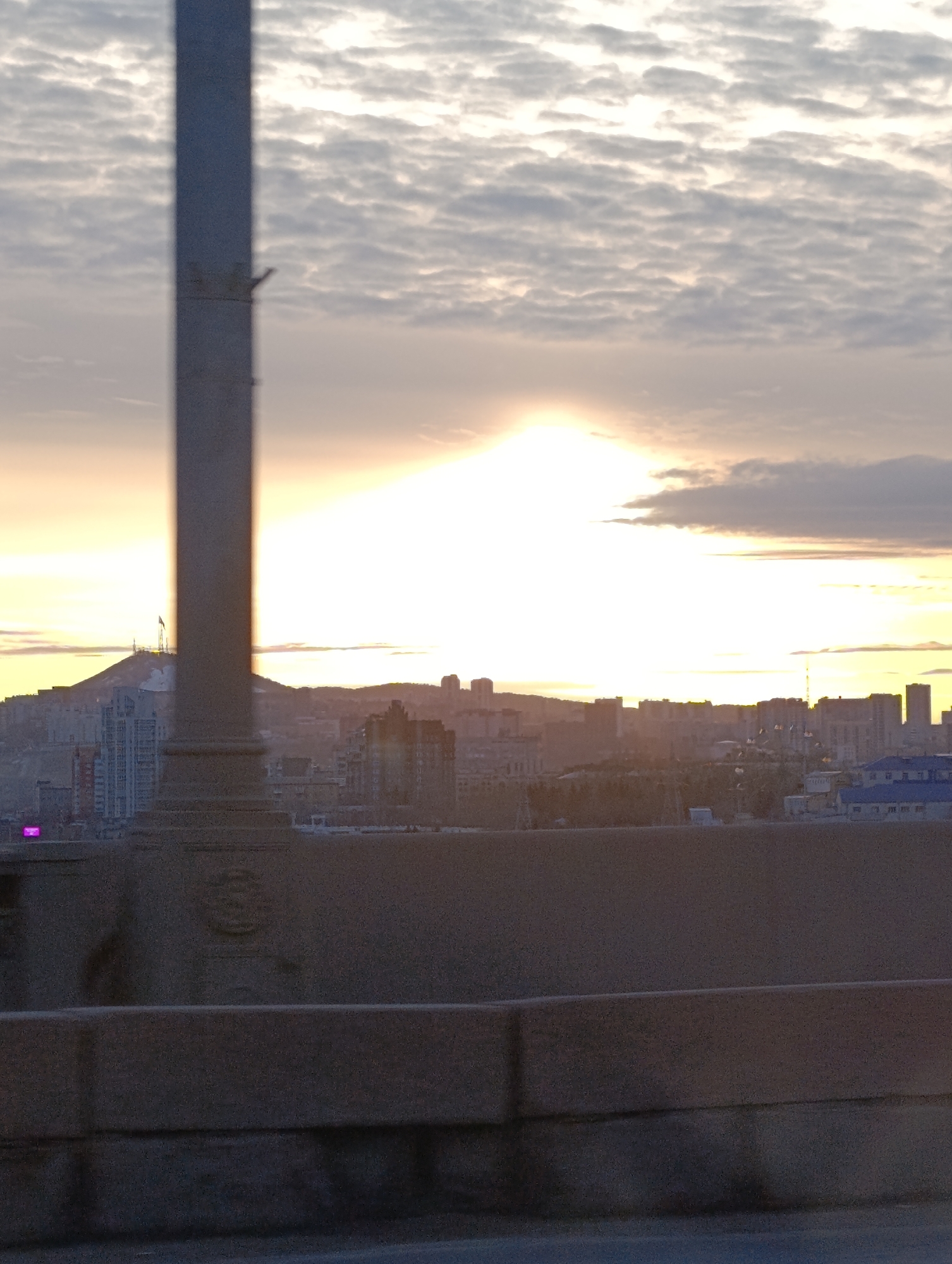 The image shows a cityscape at sunrise or sunset. The foreground features a concrete barrier, likely from a bridge or elevated road. To the left of the image is a blurred vertical structure, possibly a lamppost or pillar. Beyond the barrier, a city skyline is visible, with buildings of varying heights silhouetted against the bright light of the rising or setting sun. A prominent hill with a structure on top can be seen in the distance to the left. The sky is filled with clouds, some reflecting the warm light of the sun.
Based on the skyline and the hill with the structure on top, it is likely a view of Krasnoyarsk, Russia. The prominent hill is likely Karaulnaya Mountain, known for its chapel and communications tower.