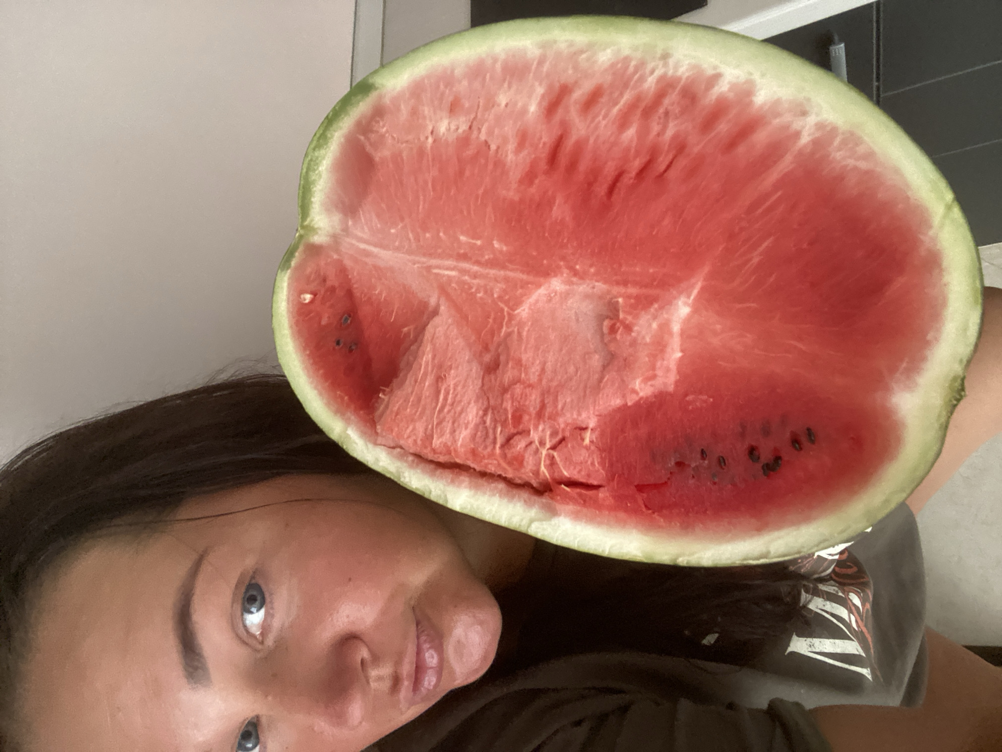 A woman with brown hair and blue eyes holds a large slice of watermelon in front of her face. Her mouth is slightly open, and she is looking towards the camera. The watermelon is vibrant pink with some visible seeds. The background is out of focus, showing a plain wall. There is no visible indication of the location being Sochi, Russia.