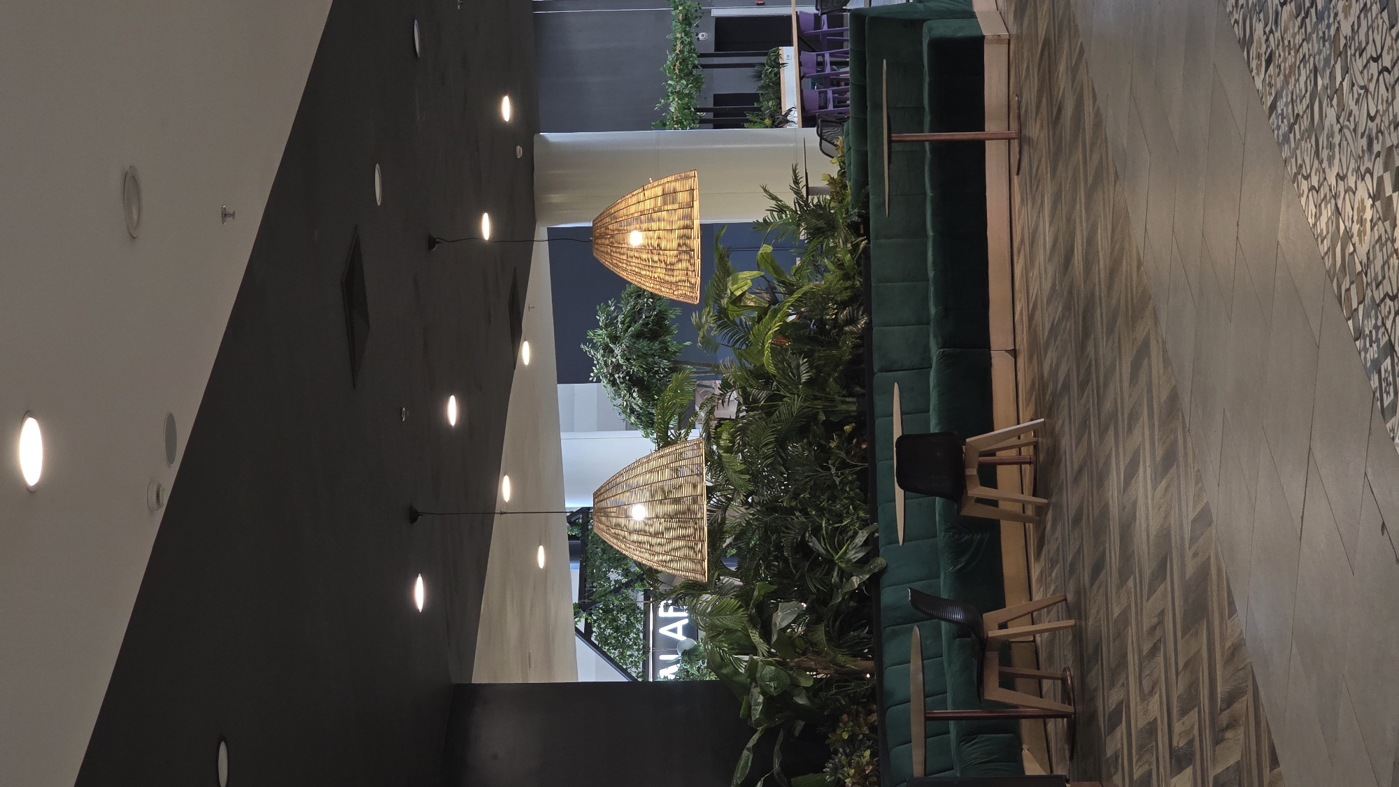 The image shows an interior space, likely a cafe or restaurant, in Stara Zagora, Bulgaria. The scene is composed of a seating area with a long, green upholstered bench along a wall filled with plants. A dark wood table sits in front of the bench, accompanied by several chairs. Two large, woven lamps hang from the ceiling above the seating area. The flooring is composed of patterned tiles in various shades of gray and brown. The ceiling consists of two distinct sections, one white with recessed lighting and the other dark gray with pendant lights. The overall impression is of a modern, nature-inspired space.