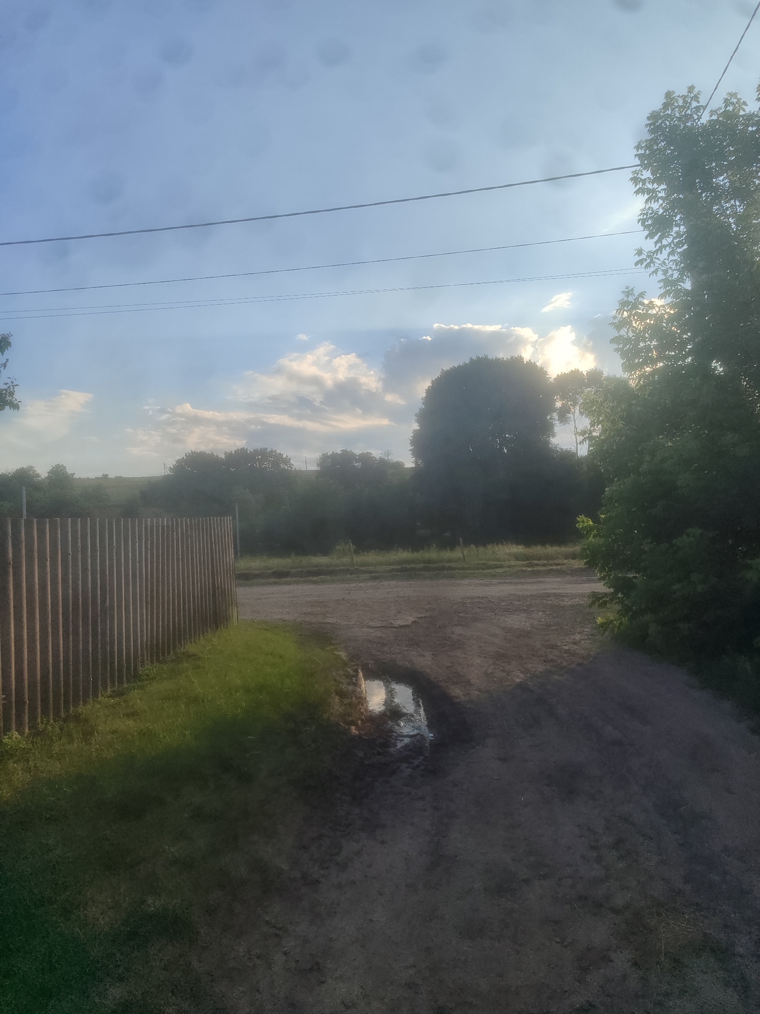 This is a daytime, outdoor shot looking across a dirt road toward a line of trees and a cloudy sky. On the left, a corrugated metal fence runs along a grassy area. The road is muddy and has a large puddle with a reflection of the sky. The trees in the background are silhouetted against the bright sky, and the sun is visible through the clouds, creating a hazy effect. The overall mood is serene and natural.