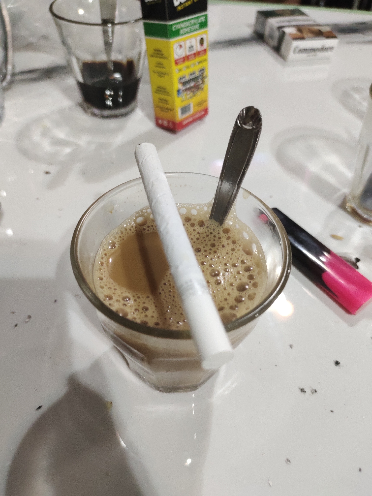 The scene is an indoor tabletop setting in Meunasahaluedrienlhoksukon Barat, Indonesia. A clear glass containing a light brown, frothy beverage, consistent with coffee or tea, is centrally positioned. A white, unfiltered cigarette is inserted diagonally into the beverage, with its filter end submerged, while a silver metal spoon rests upright beside it. To the upper left, a second clear glass, partially visible, contains a dark liquid and a spoon. In the upper right background, a yellow and green rectangular product box, partially labeled, stands near a visible pack of "Commodore" cigarettes. A black and pink object, resembling a marker or lighter, lies to the right of the central glass. The white tabletop surface exhibits scattered dark particulate matter and reflections from an unseen light source. No individuals are present in the image.
