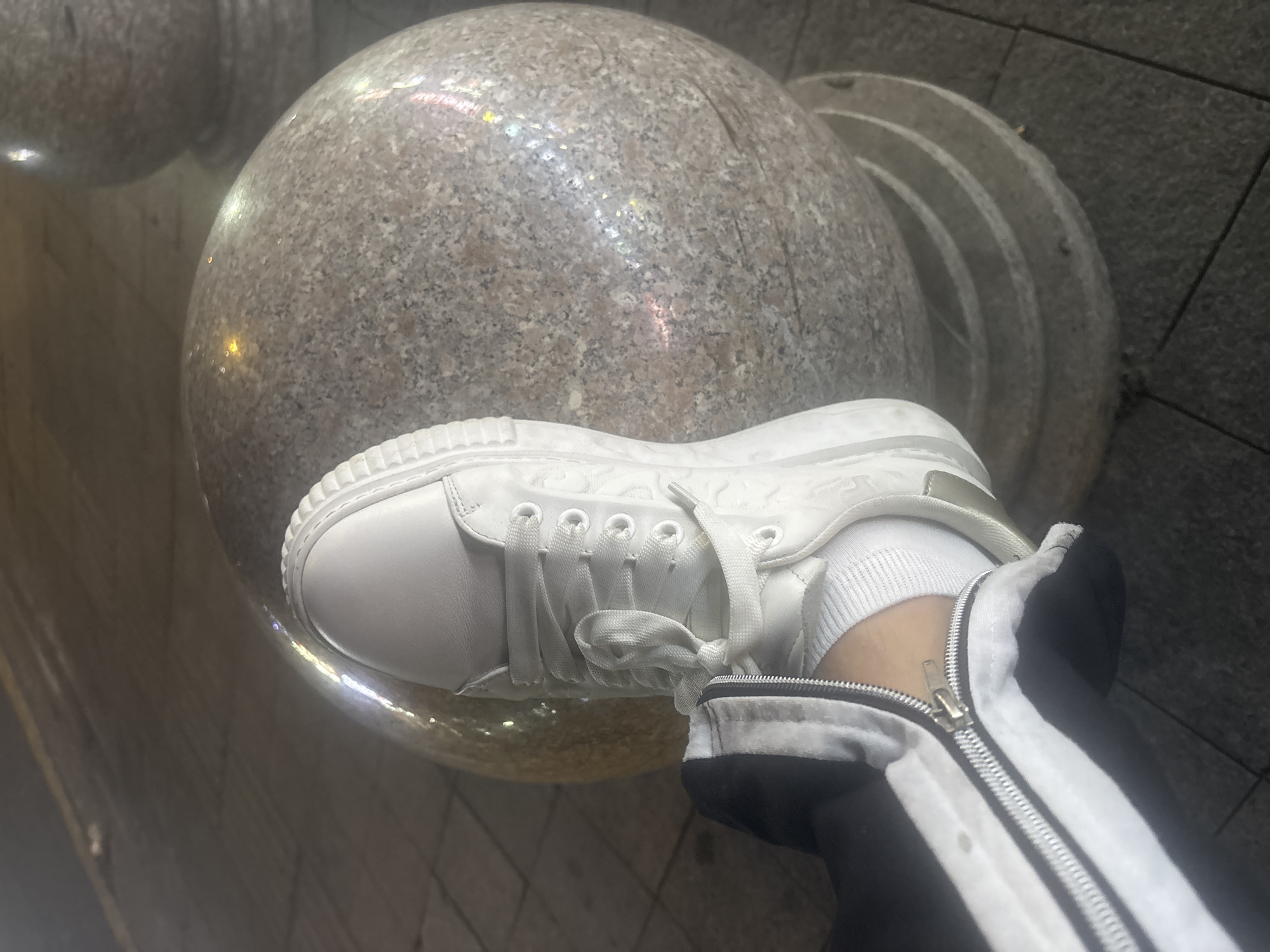 A person's white sneaker with white laces rests on a large, grey, speckled sphere, possibly a decorative boulder. The shoe is laced up, and a white sock is visible. The person is wearing dark pants with a white stripe and a zipper that is partially open, revealing the ankle. The scene appears to be outdoors, on a paved surface, possibly a sidewalk or plaza, with the background showing a slightly blurred street or public space at night, with reflections from streetlights on the ground and the sphere. The location is in Suzhou Industrial Park, China.