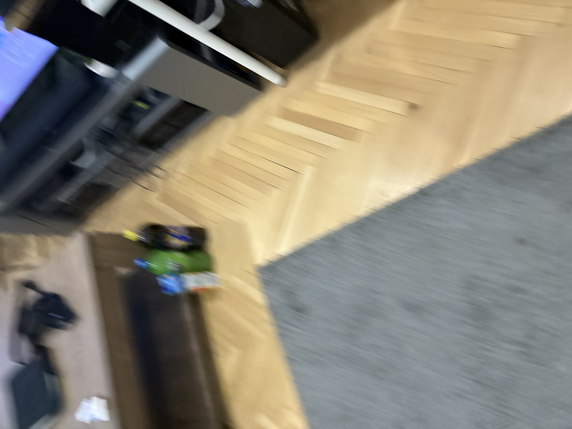 The image shows a blurred overhead view of a section of a room. Visible are:
* Part of a TV stand and TV in the upper left corner.
* Wood flooring.
* A gray rug in the lower right corner.
* A black object resembling a vacuum cleaner in the lower left.
* Several bottles and packets on the floor near the rug.
There is no information to determine the location of this scene to be in Lovech, Bulgaria. No people are visible. The actions and interactions are not evident in the blur of the image.