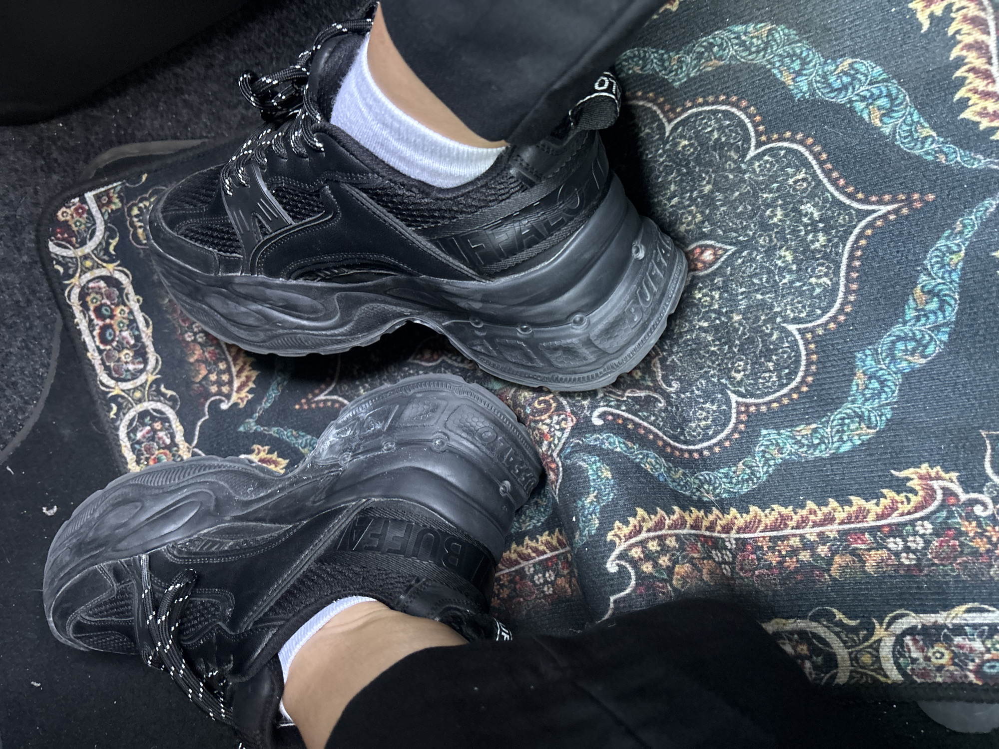 A high-angle, close-up shot captures a person's legs and feet, adorned in black chunky sneakers, resting on a decorative floor mat. The person wears black trousers and white ankle socks, with black sneakers featuring thick, sculptural soles and black-and-white patterned laces. The brand name "BUFFALO" is prominently visible in raised black lettering on the side of both sneakers' uppers, near the ankle, and also on the side of the soles. A small black loop with "LO" (part of BUFFALO) is visible at the back of the right shoe.
The feet are positioned on a dark, patterned mat. This mat features intricate, oriental-style motifs in shades of dark blue, teal, gold, red, and beige, against a dark background. Beneath the patterned mat, a dark grey, carpeted surface is visible, suggesting the interior of a vehicle, likely a car. No specific activity or time of day is discernible. The scene is located in Budva, Montenegro.