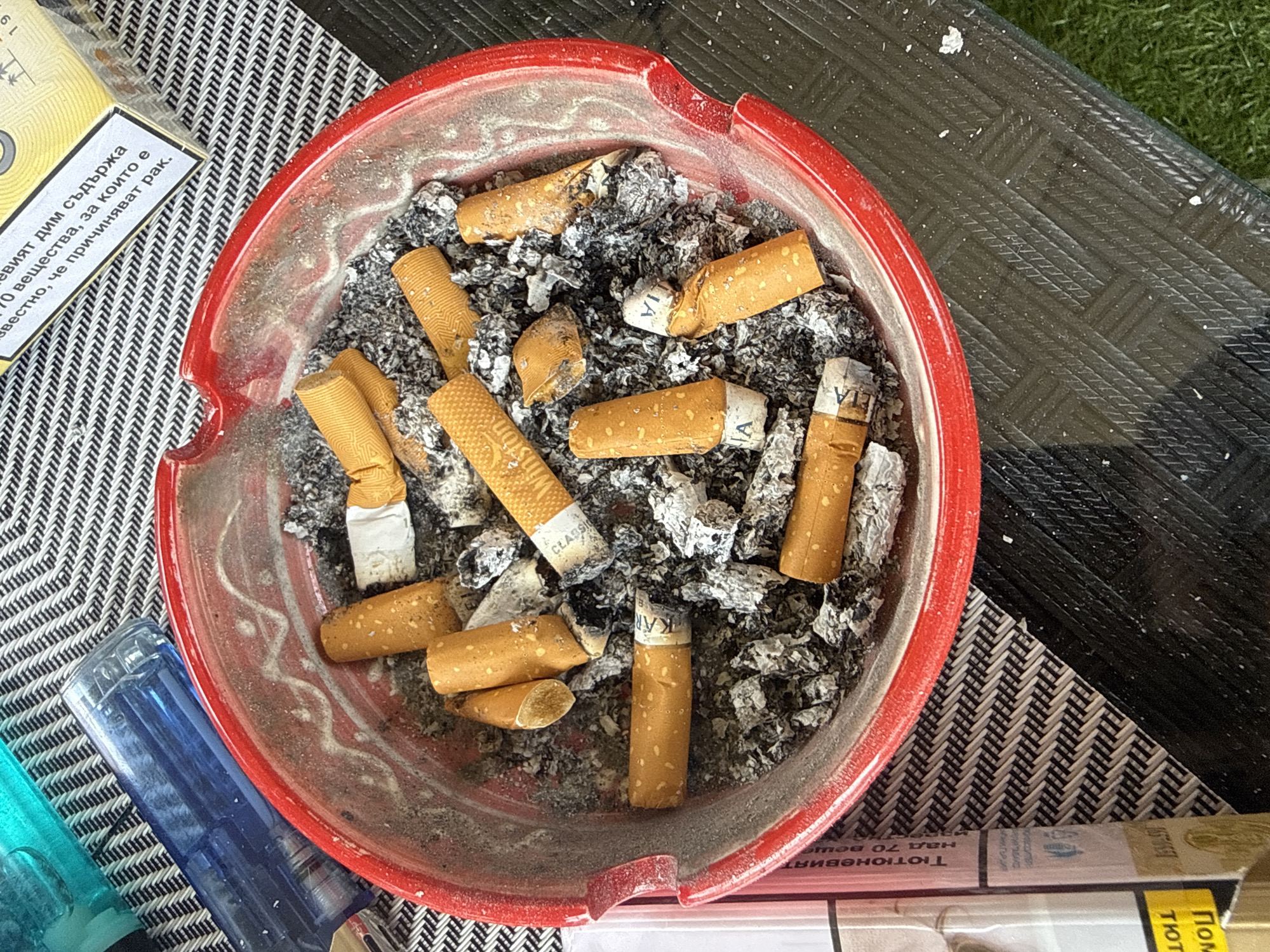 The image depicts an overflowing red ashtray filled with numerous cigarette butts and ash. Several cigarette butts are whole, while others are broken or partially consumed. Some butts have visible branding, including "Winston" and "CLASSIC." A blue lighter is positioned to the left of the ashtray, atop a black and white striped mat. In the upper right corner, a portion of a cigarette pack is visible, featuring text in a foreign language, likely warning labels related to smoking. The ashtray and surrounding surfaces appear dusty and grimy, suggesting recent use. The background consists of a dark, textured surface, possibly a table, with a hint of green grass in the lower right corner. This scene is suggestive of an outdoor setting, possibly a patio or garden area. There are no people or animals present in the image. The image does not provide any specific information about the location in the city of Colchester, United Kingdom.