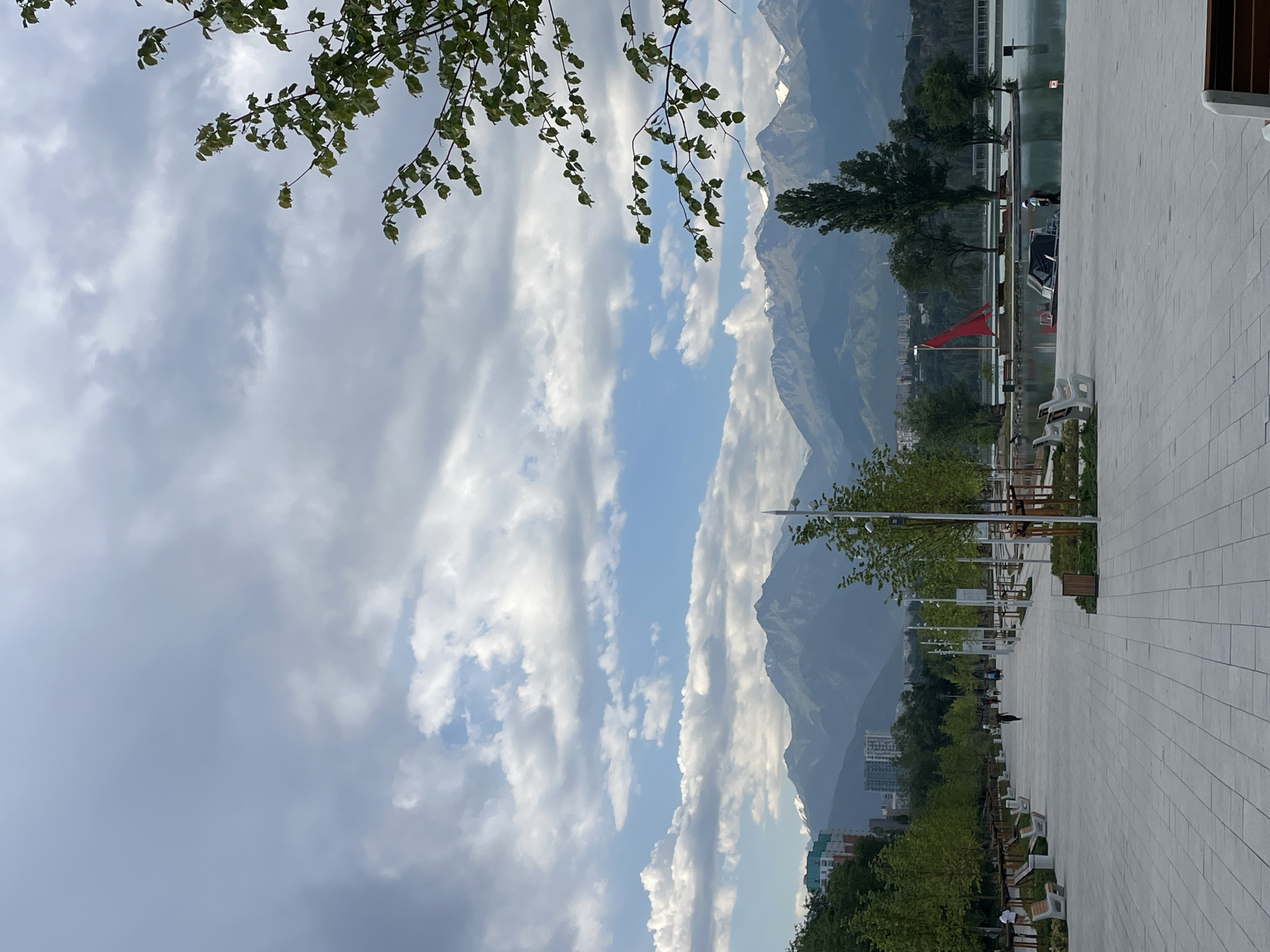 The image depicts a view of the First President's Park in Almaty, Kazakhstan. The park features a wide, paved walkway lined with trees and benches, leading towards a body of water. In the background, a range of majestic mountains dominates the skyline under a sky filled with dramatic clouds. A red sail, possibly from a small boat or decorative element, is visible on the water. A few people are present, walking along the path and near the water's edge. The overall scene conveys a sense of serene urban nature.