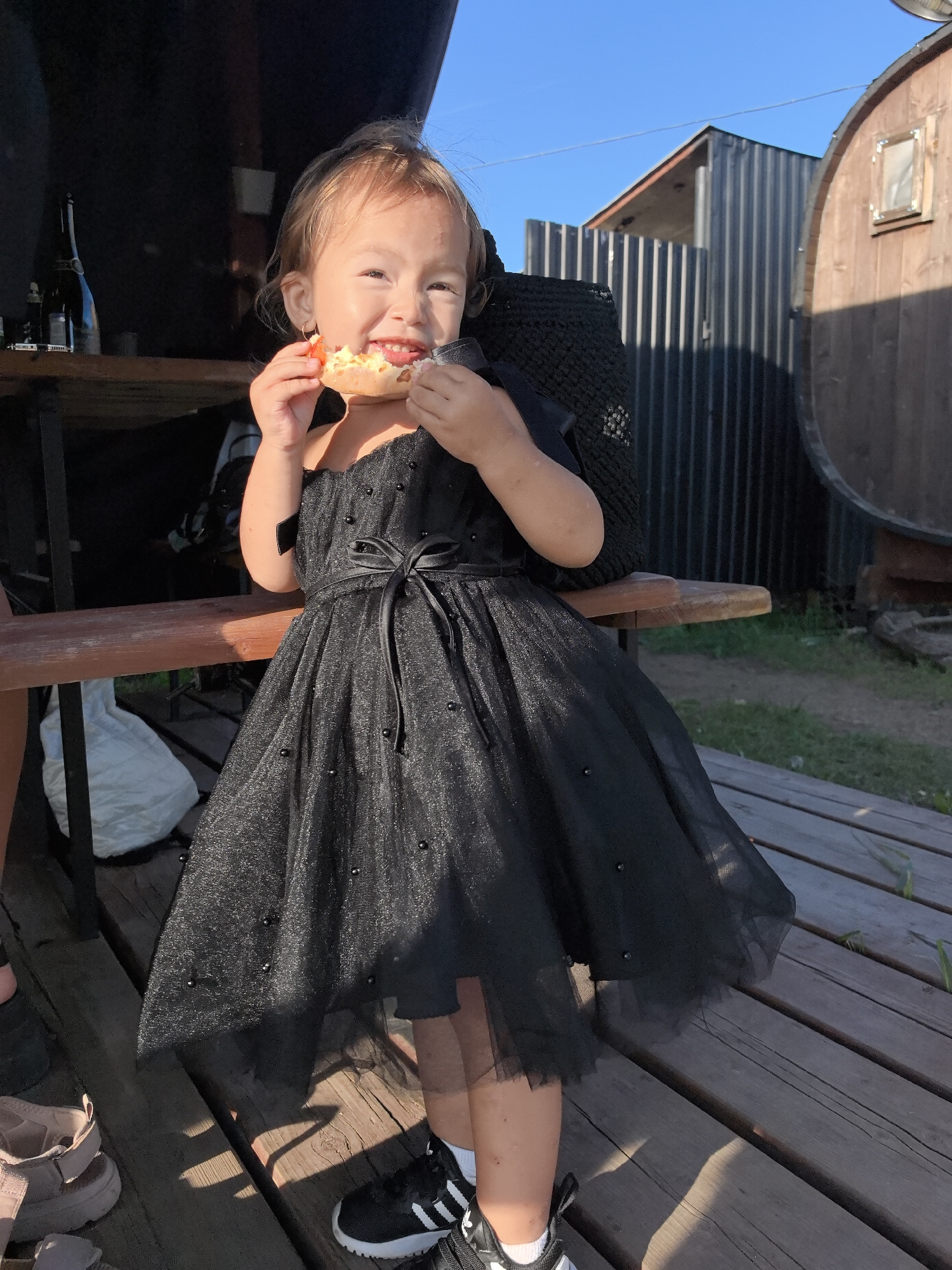 A young child, wearing a black tulle dress adorned with pearls and a black ribbon belt, is pictured outdoors on a sunny day. The child is smiling and holding a piece of food, possibly a pastry or bread, up to their mouth, taking a bite. The child is standing on a wooden deck, with a wooden bench in the foreground. In the background, there are various structures including a corrugated metal building and a large wooden barrel. The setting appears to be a rural or semi-rural environment, potentially in the city of Olkhovka, Russia, given the presence of traditional structures like wooden barrels and wooden decks commonly found in such areas. The overall atmosphere is casual and bright, with sunlight casting shadows on the wooden planks.