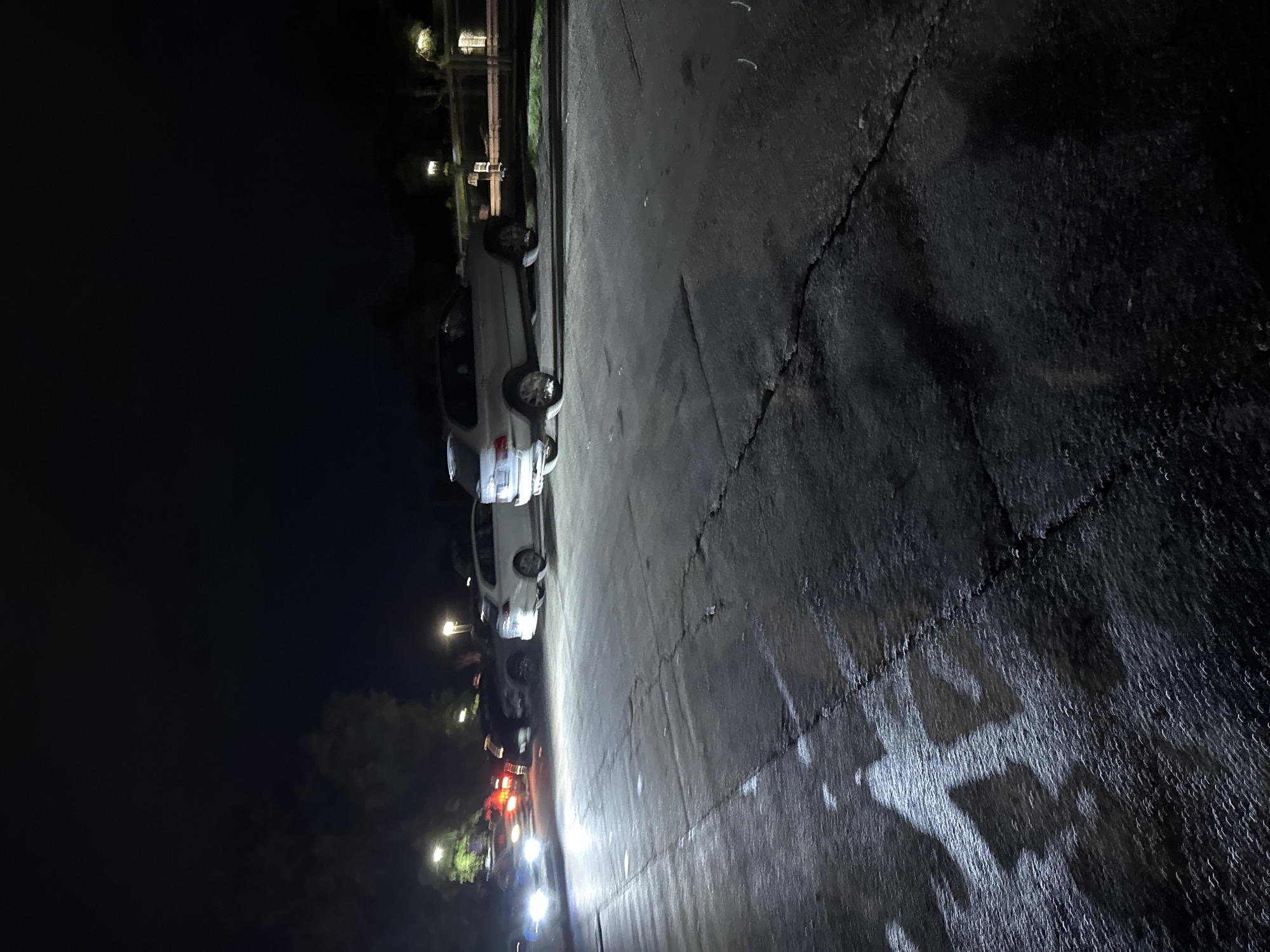 The image depicts a nighttime street scene with cars parked along the left side. The street surface appears to be wet, reflecting light from the vehicles and streetlights. The background is dark, suggesting it is late at night with minimal ambient light. The location is unknown.