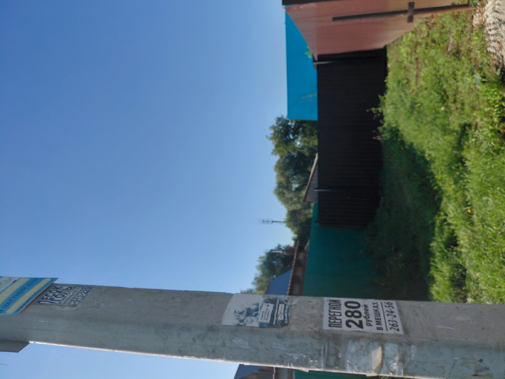 The image shows a concrete utility pole with multiple advertisements. One prominent advertisement is a rectangular sticker with black text on a white background. The text reads "ПЕРЕГНОЙ 280 рублей В МЕШКАХ 263-24-56". Above this, there is another sticker with blue and yellow text. The pole is situated outdoors, with a clear blue sky in the background. Beyond the pole, there is a green fence and some trees. To the right, a portion of a red-brown fence is visible. The image appears to be taken on a sunny day. The advertisement suggests the sale of humus (перегной) in bags for 280 rubles, with a contact phone number 263-24-56. The presence of a utility pole with advertisements is common in urban and suburban areas. The style of the advertisements and the language used (Russian) suggest a location within Russia. Without further context or identifying landmarks, it is not possible to definitively determine the specific location within Novosibirsk, Russia.